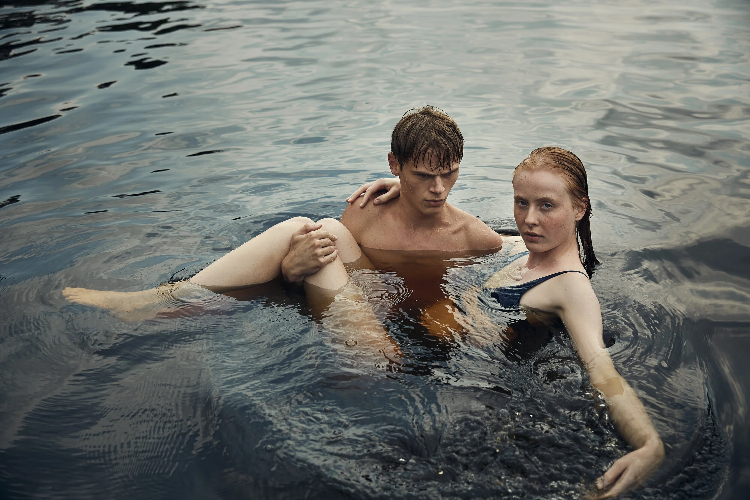 Ein Mann und eine Frau schwimmen in einem Gewässer. Der Mann hält die Beine der Frau, beide sehen ernst in die Kamera. Die Umgebung ist von Wasser umgeben.