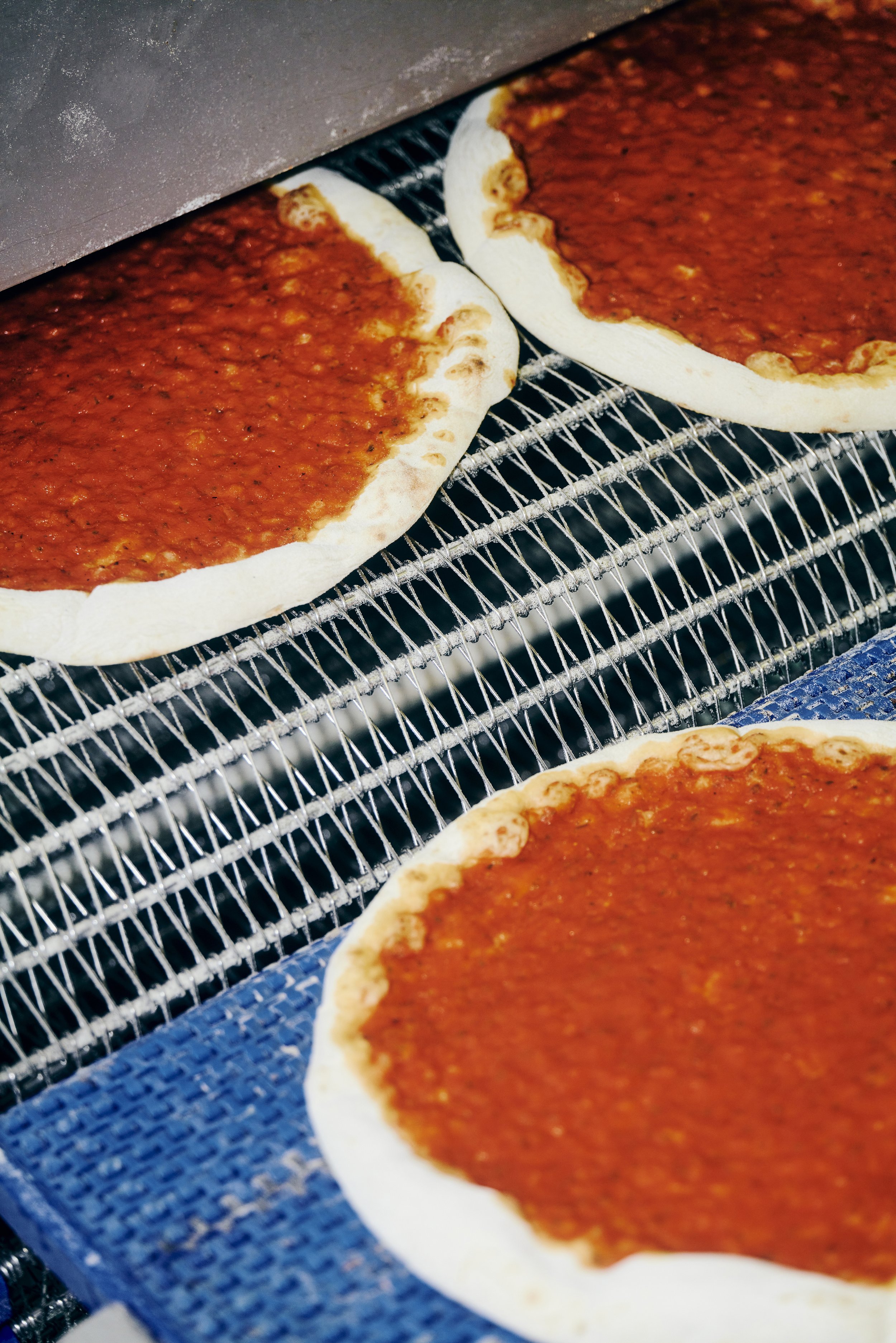 Pizzen mit Tomatensauce auf einem Förderband in einem Ofen.