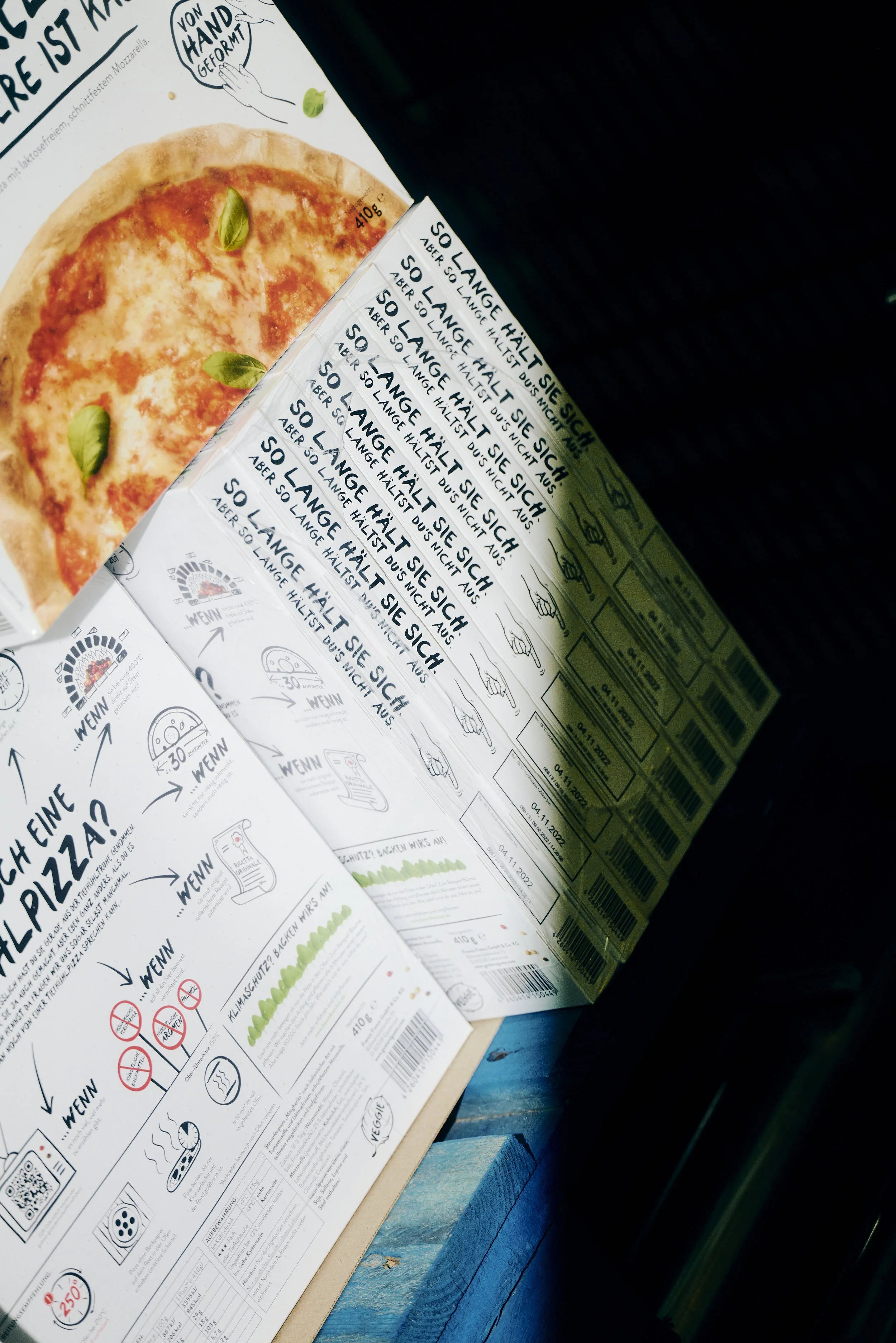 Verpackung von Tiefkühlpizzen mit grafischen Anleitungen auf der Rückseite. Die Vorderseite zeigt ein Bild einer Pizza mit Basilikumblättern. Mehrere Informationen und Illustrationen sind sichtbar, die den Gebrauch erklären.