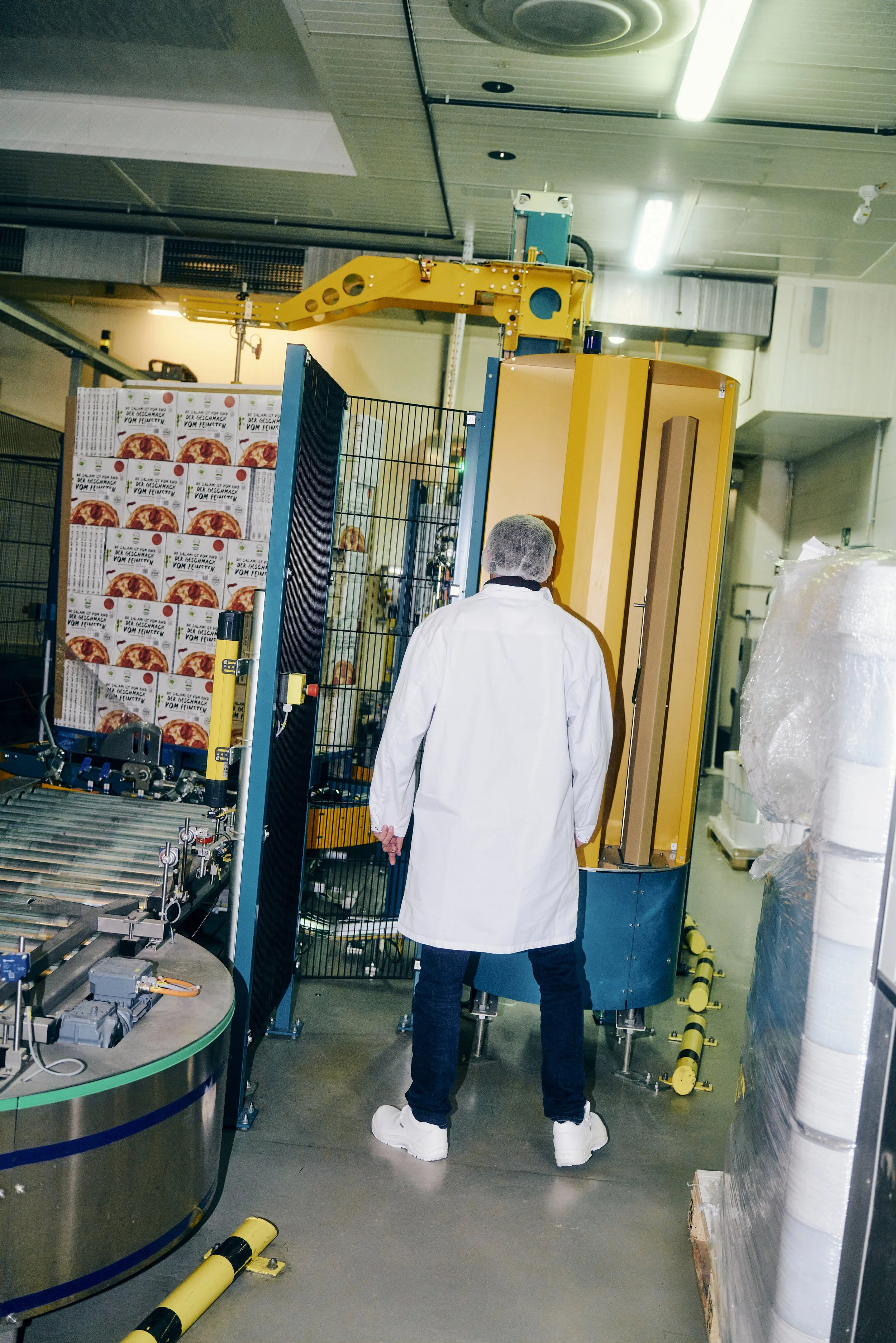 Person in weißem Mantel steht in einer Fabrik neben Maschinen und gestapelten Pizzapackungen.