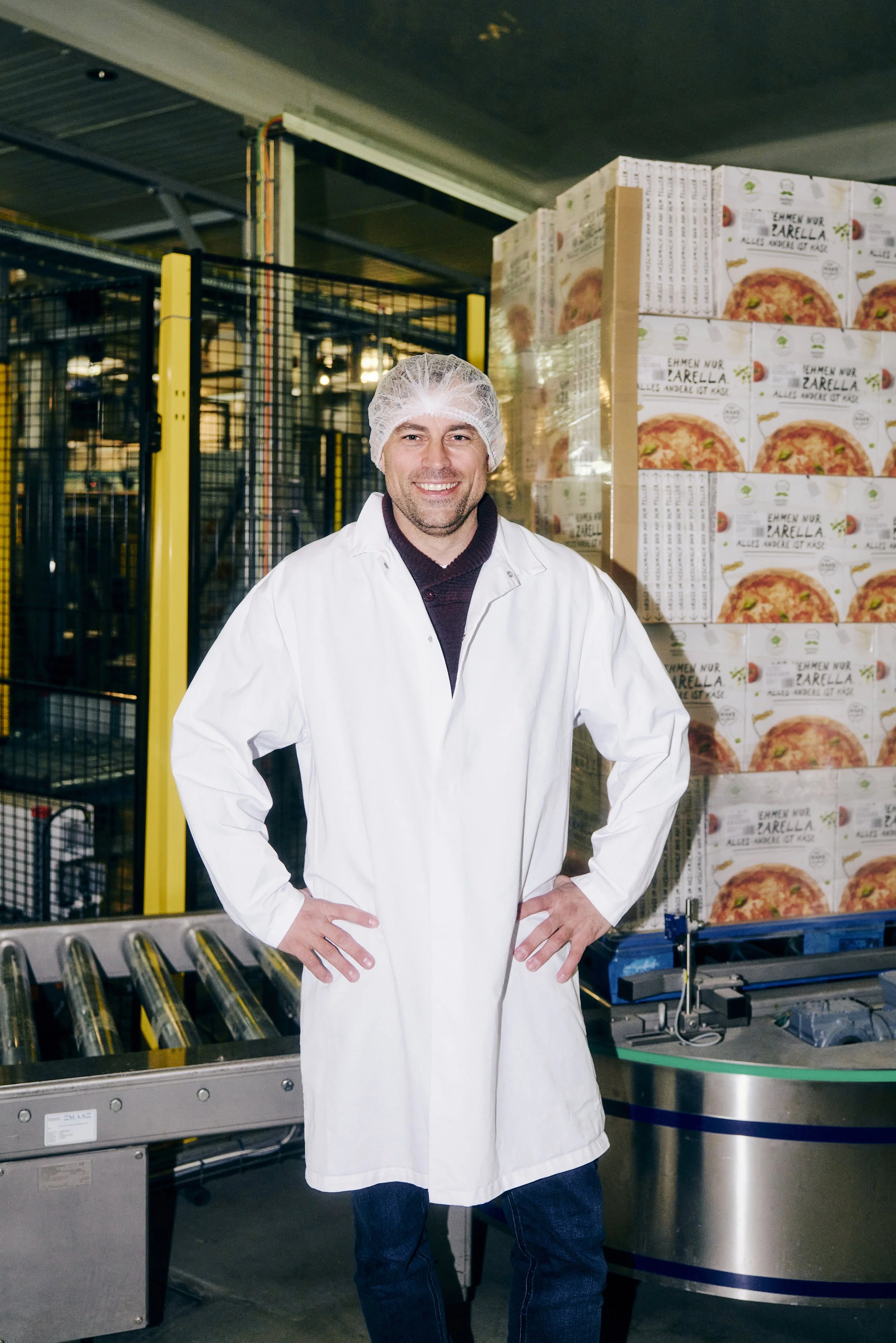 Mann in Schutzkleidung steht in einer Fabrik mit gestapelten Lebensmittelkartons im Hintergrund.