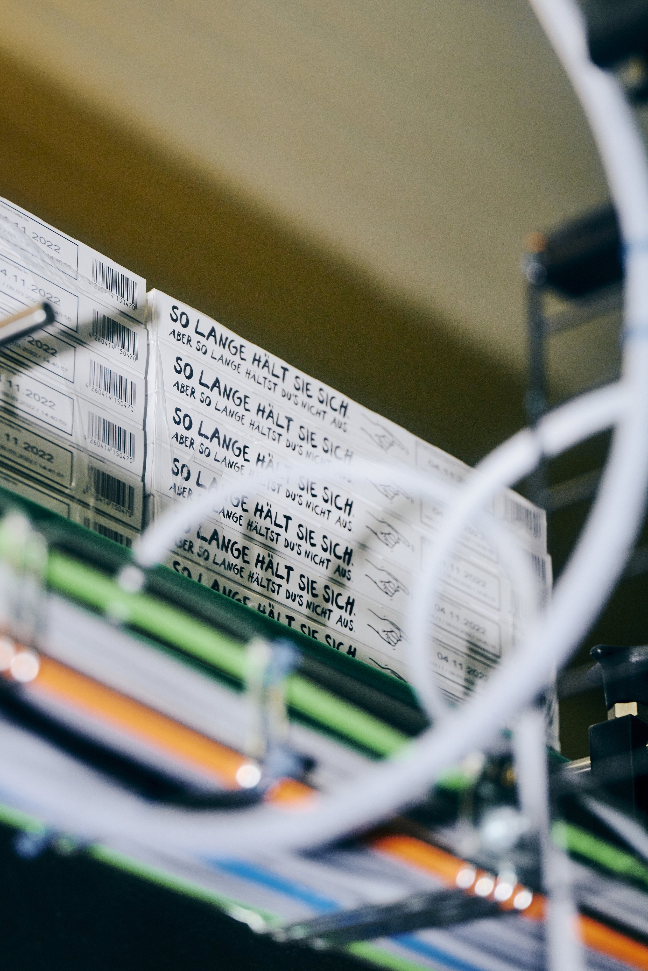 Nahaufnahme von beschrifteten Verpackungskartons mit dem Text "SO LANGE HÄLT SIE SICH" und weiteren Details, umgeben von Kabeln im Vordergrund.