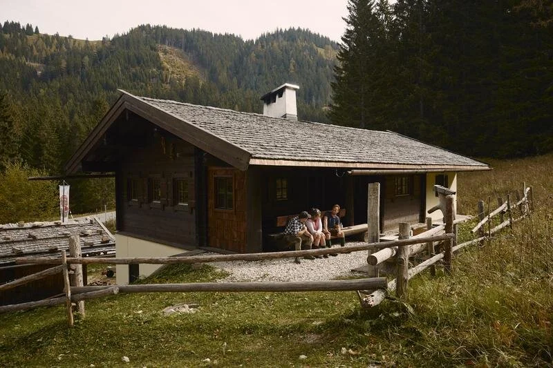Traditionelle Berghütte mit Holzdach und Holzzaun inmitten einer waldreichen alpinen Landschaft, vier Personen sitzen auf einer Bank vor der Hütte.
