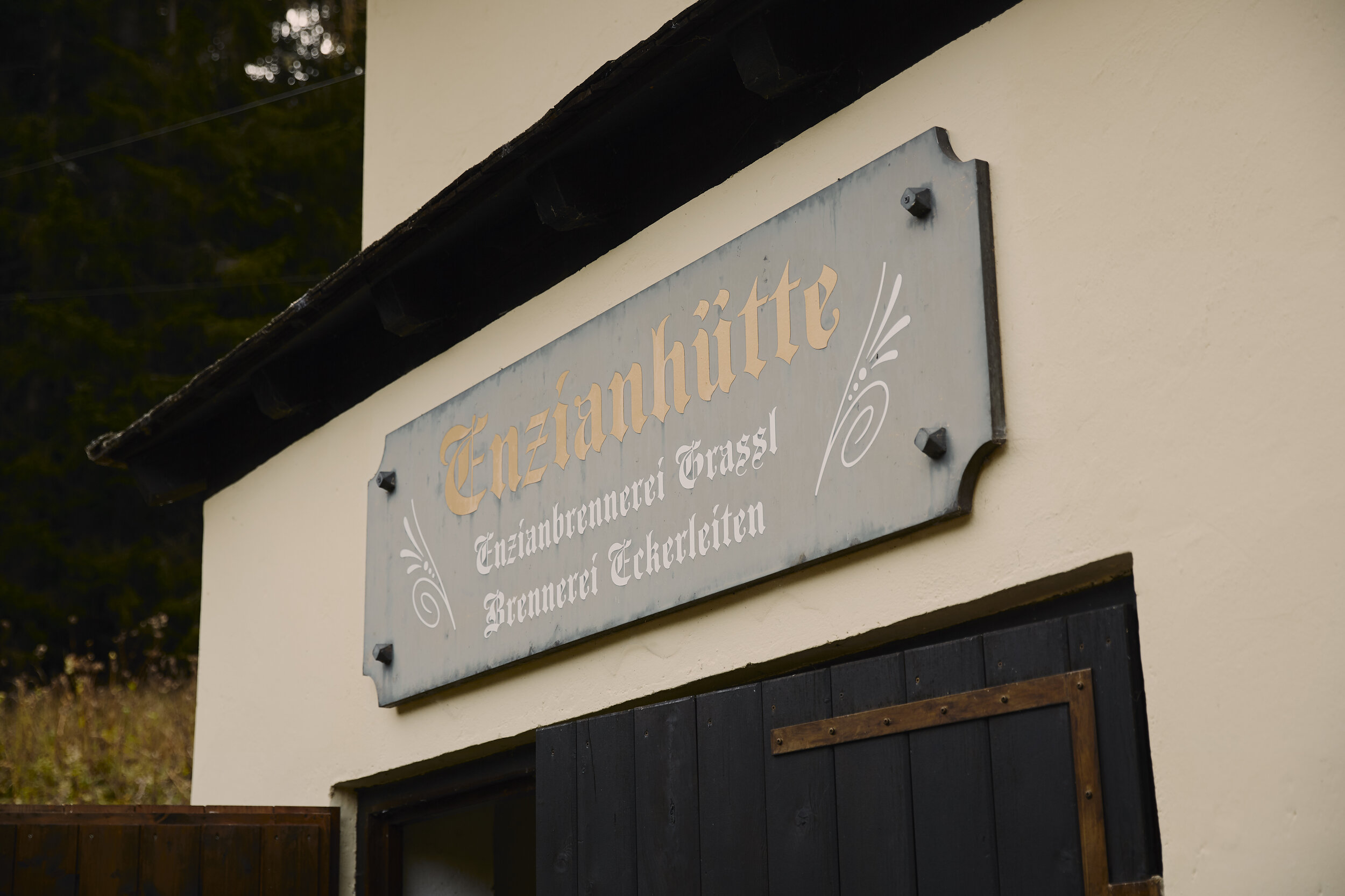 Gebäude mit einem Schild "Enzianhütte" und "Enzianbrennerei Grassl", mit dekorativen Elementen und dahinterliegendem Wald.