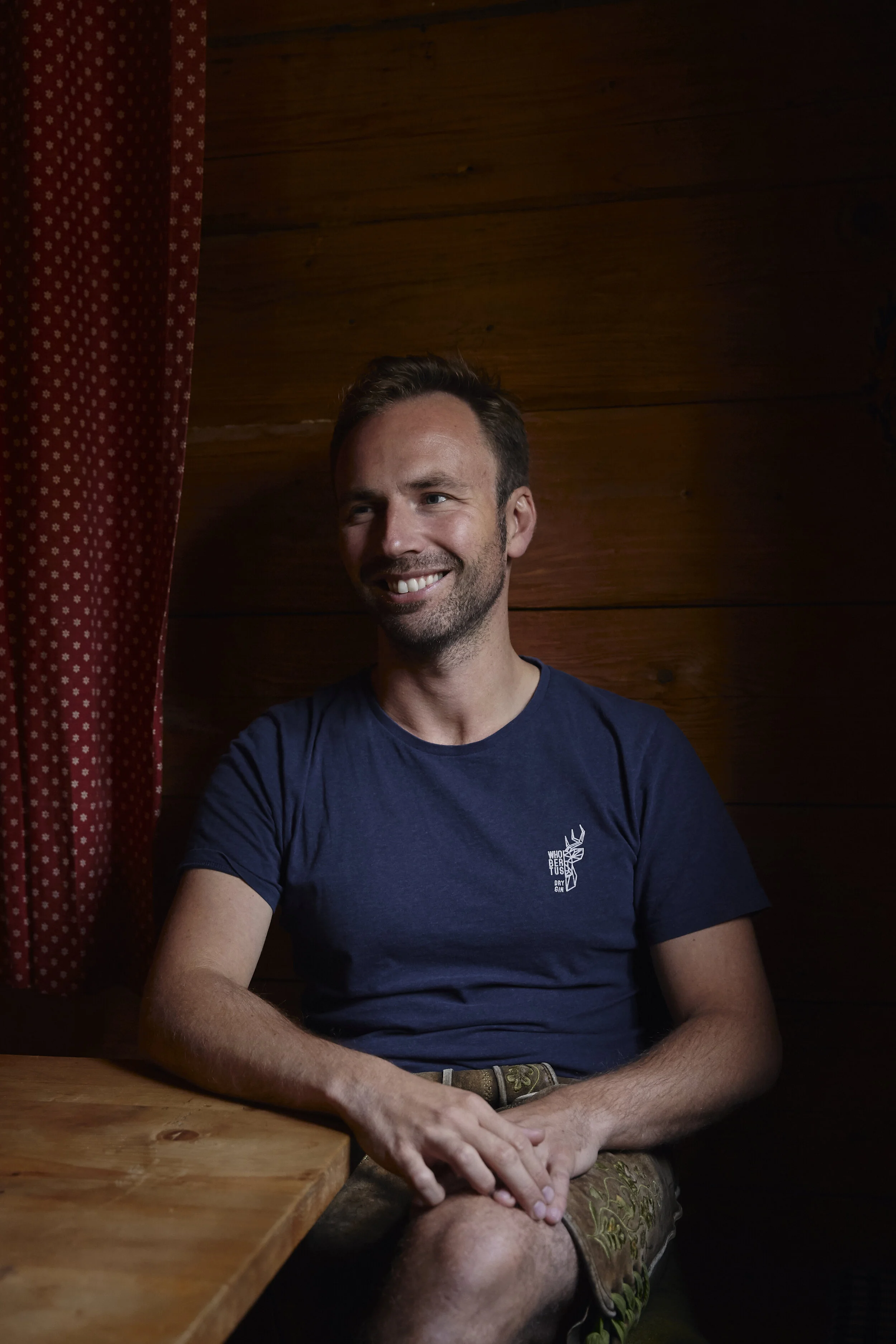 Ein Mann sitzt in einem Raum mit Holzvertäfelung, trägt ein dunkelblaues T-Shirt und lächelt. Neben ihm ist ein rot-weiß gepunkteter Vorhang sichtbar.