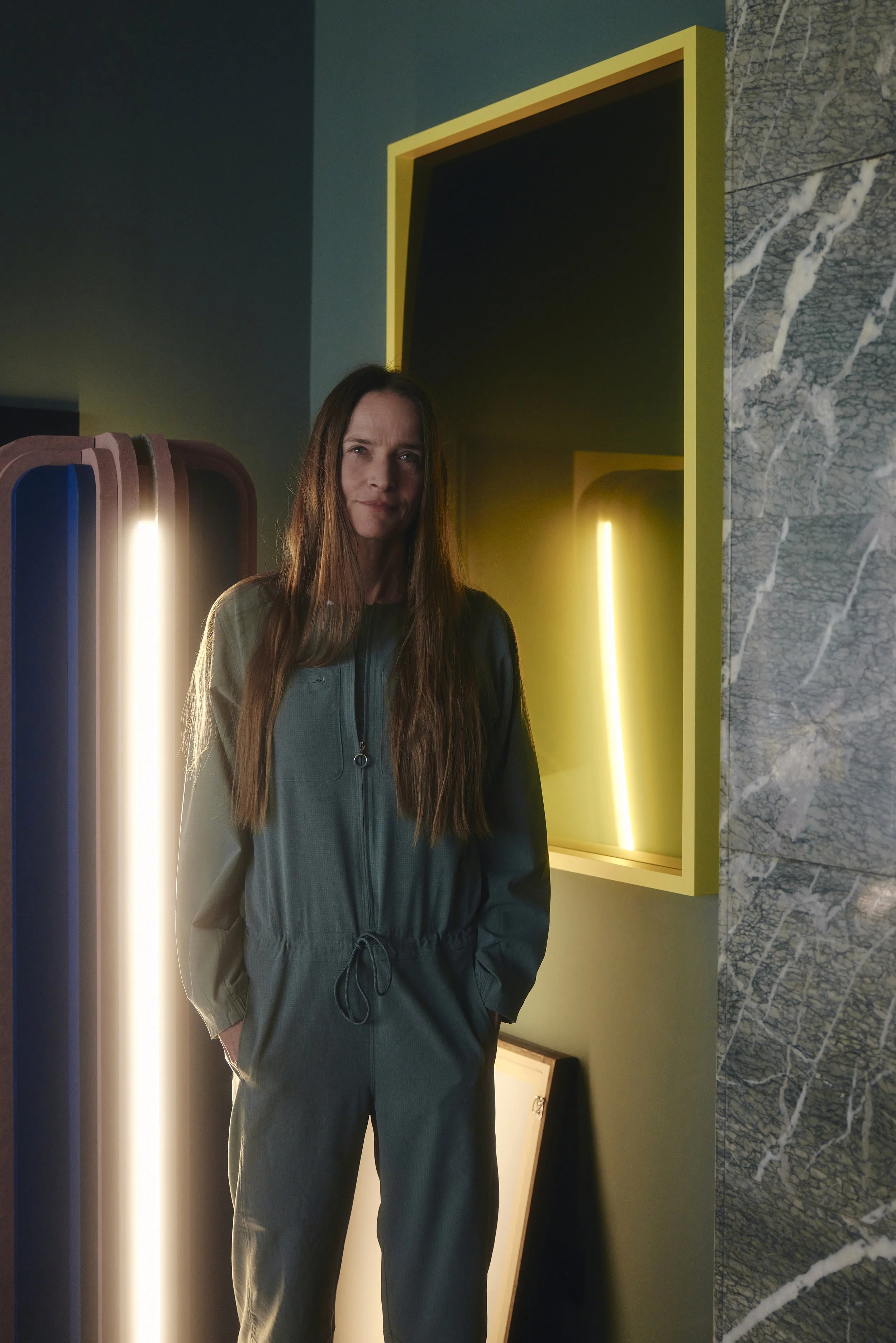 Marie Aigner in einem grünen Overall steht in einem modern gestalteten Raum mit einem leuchtenden Kunstwerk im Hintergrund.