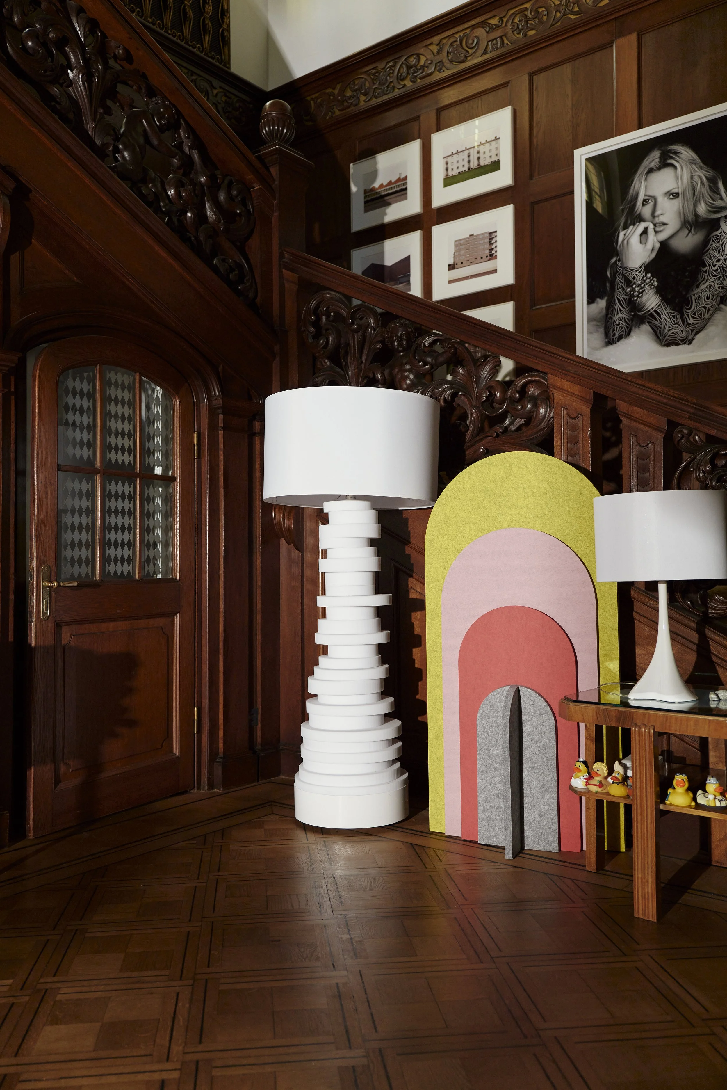 Innenraum mit Holztreppe, dekorative Lampen, bunte Deko-Elemente, Gummienten auf Ablage, gerahmte Fotos und Porträt an der Wand.