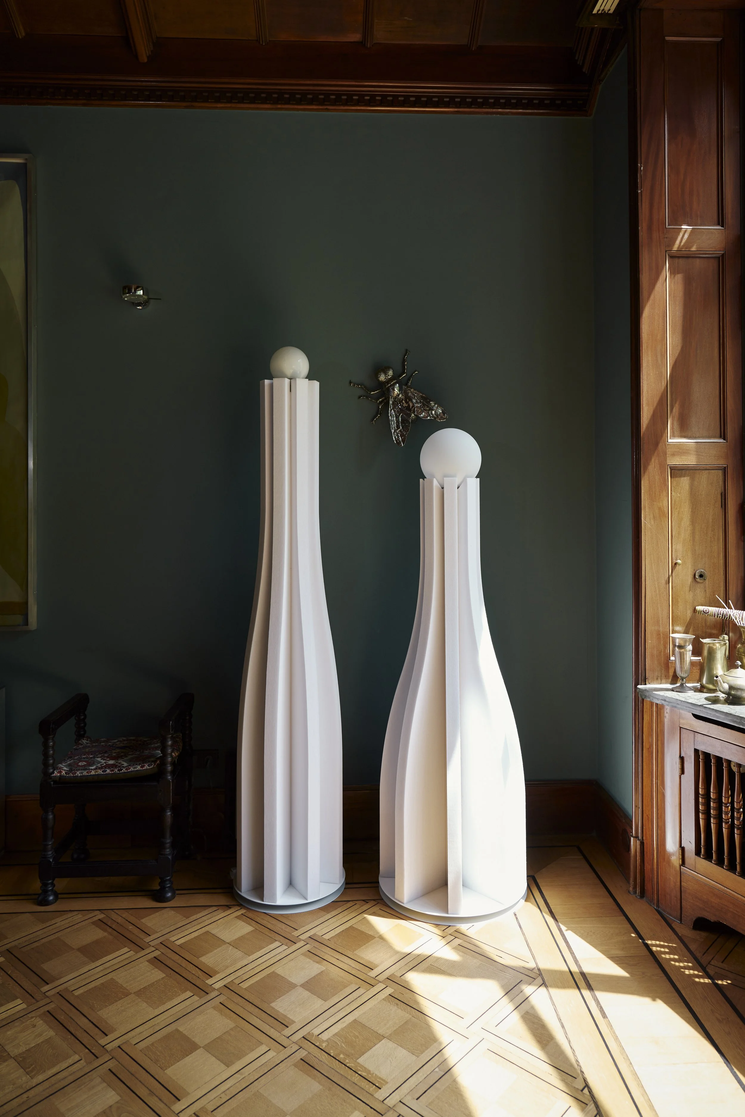 Zwei hohe, weiße, wellenförmige Skulpturen mit Kugeln oben auf einem Holzboden in einem Raum mit grüner Wand und Holzverkleidung. Links steht ein kleiner Stuhl und an der Wand hängt eine metallische Fliegenstatue.