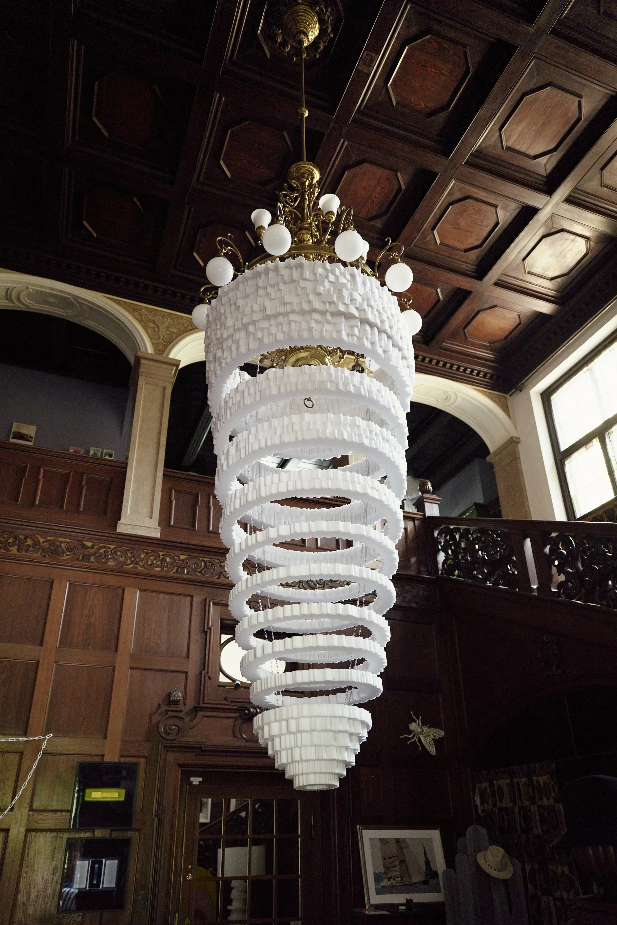 Auf dem Bild ist ein großer Kronleuchter aus Metall und weißen Ornamenten in einem Holzraum mit verzierten Paneelen an der Decke zu sehen.