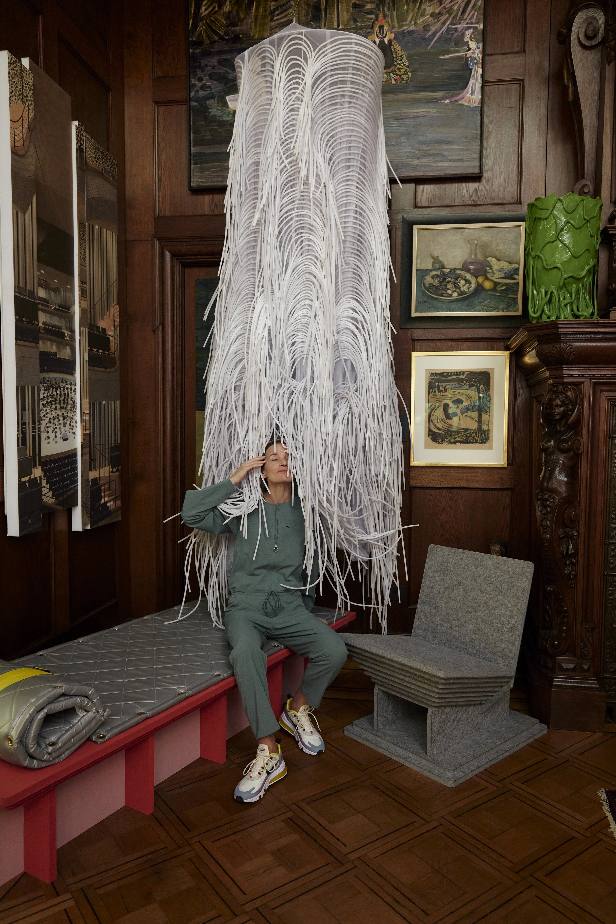 Person in einem Raum mit Holzwänden hebt einen großen, weißen, strukturreichen Gegenstand über den Kopf, der aus langen, schmalen Streifen besteht. Der Raum ist mit verschiedenen Gemälden und Kunstwerken dekoriert.