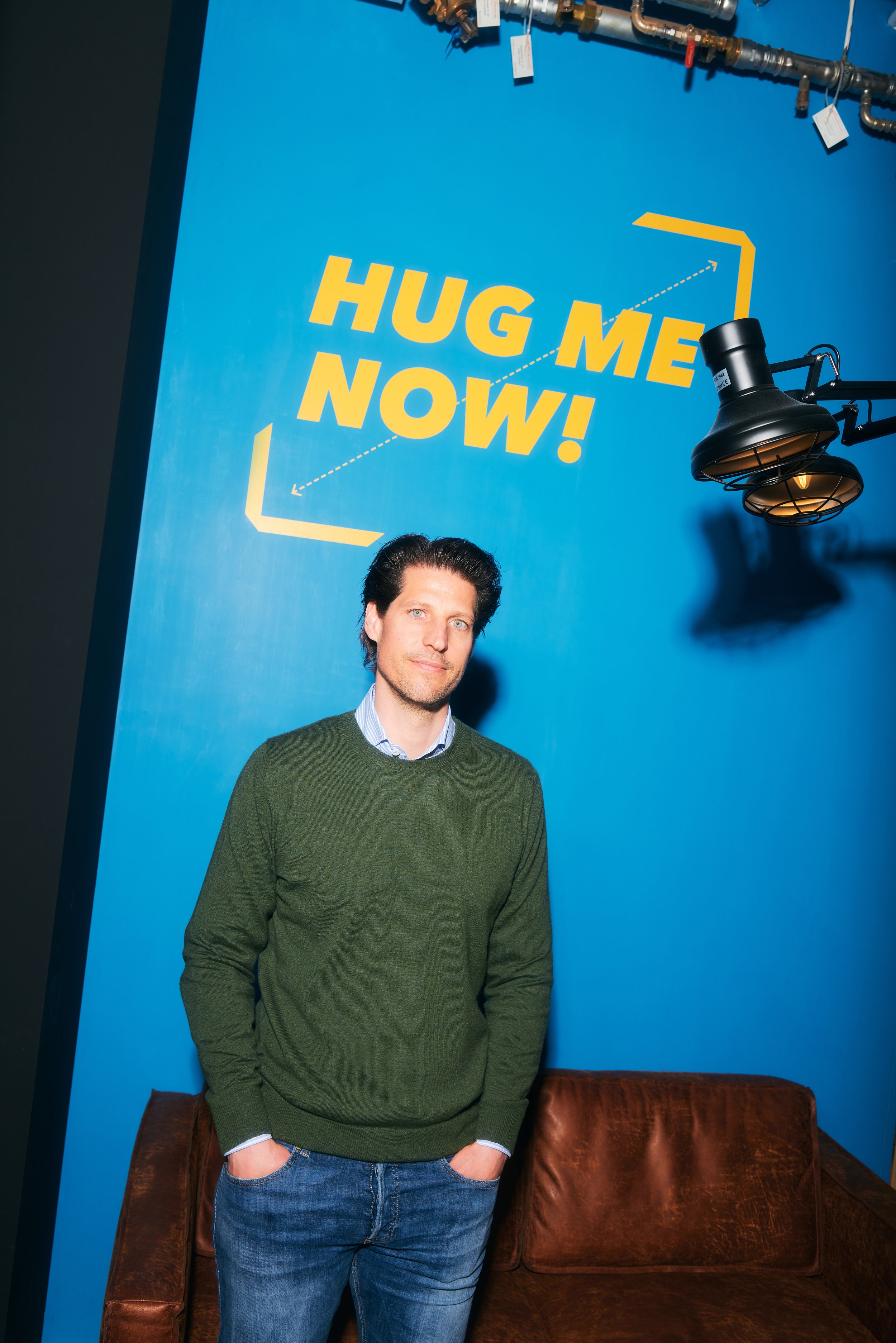 Ein Mann steht vor einer blauen Wand mit der Aufschrift 'HUG ME NOW!'. Er trägt einen grünen Pullover über einem Hemd und eine blaue Jeans. Neben ihm steht ein Ledersofa und eine Lampe ist an der Wand montiert.