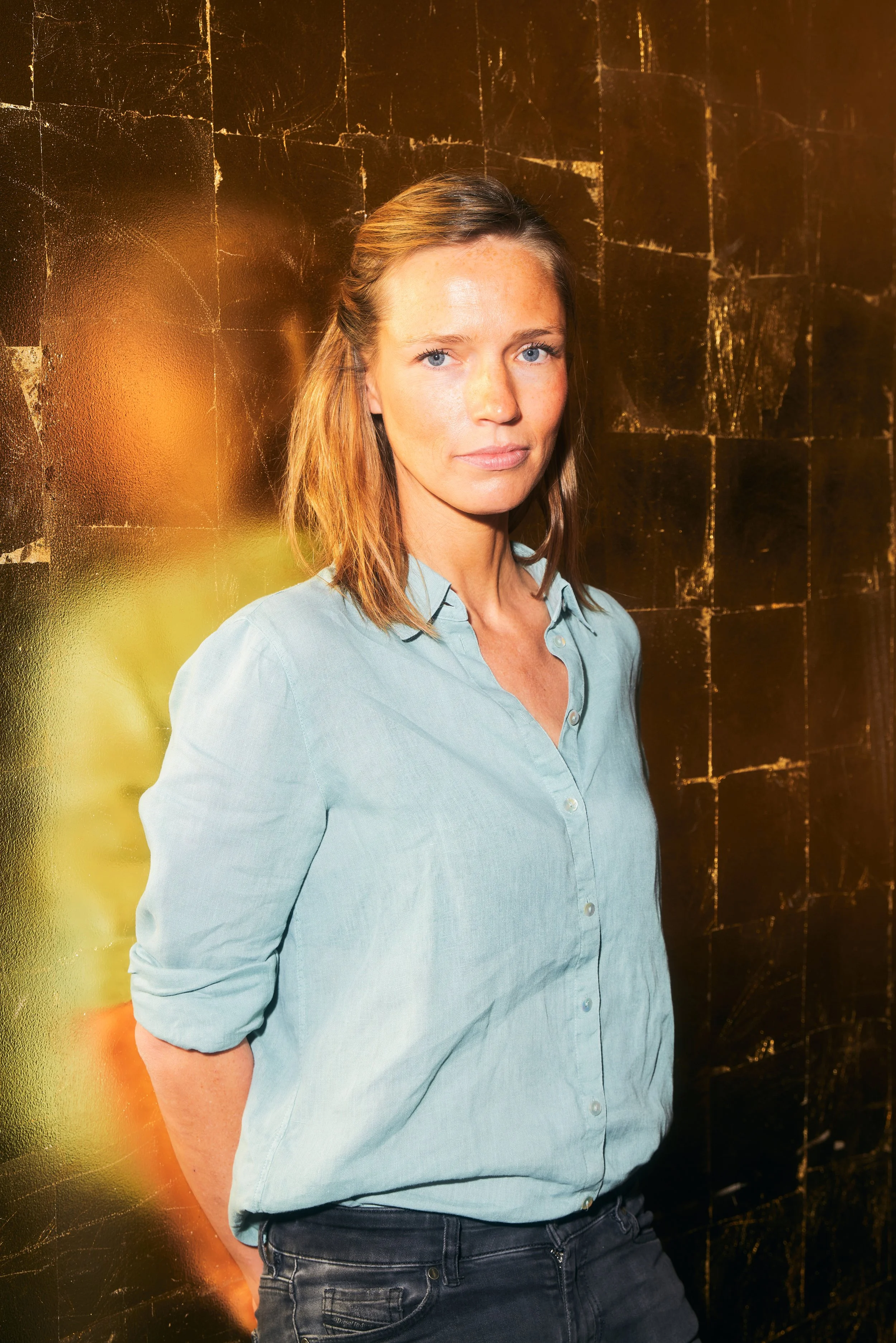 Frau vor einer goldenen Wand, trägt ein blaues Hemd.