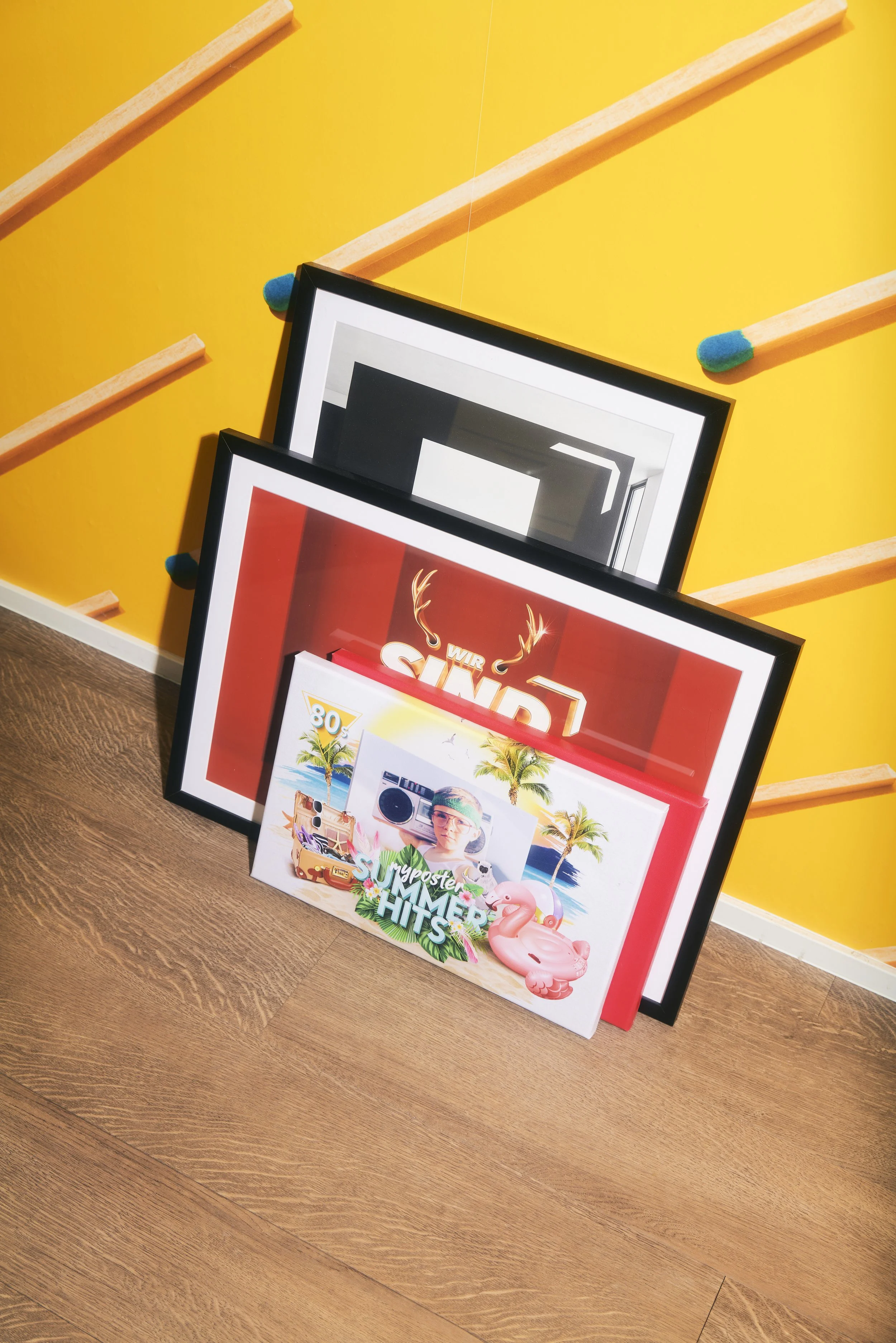 Gerahmte Poster an gelber Wand, eins mit Flamingo und "Summer Hits" Thema, auf Holzboden platziert.