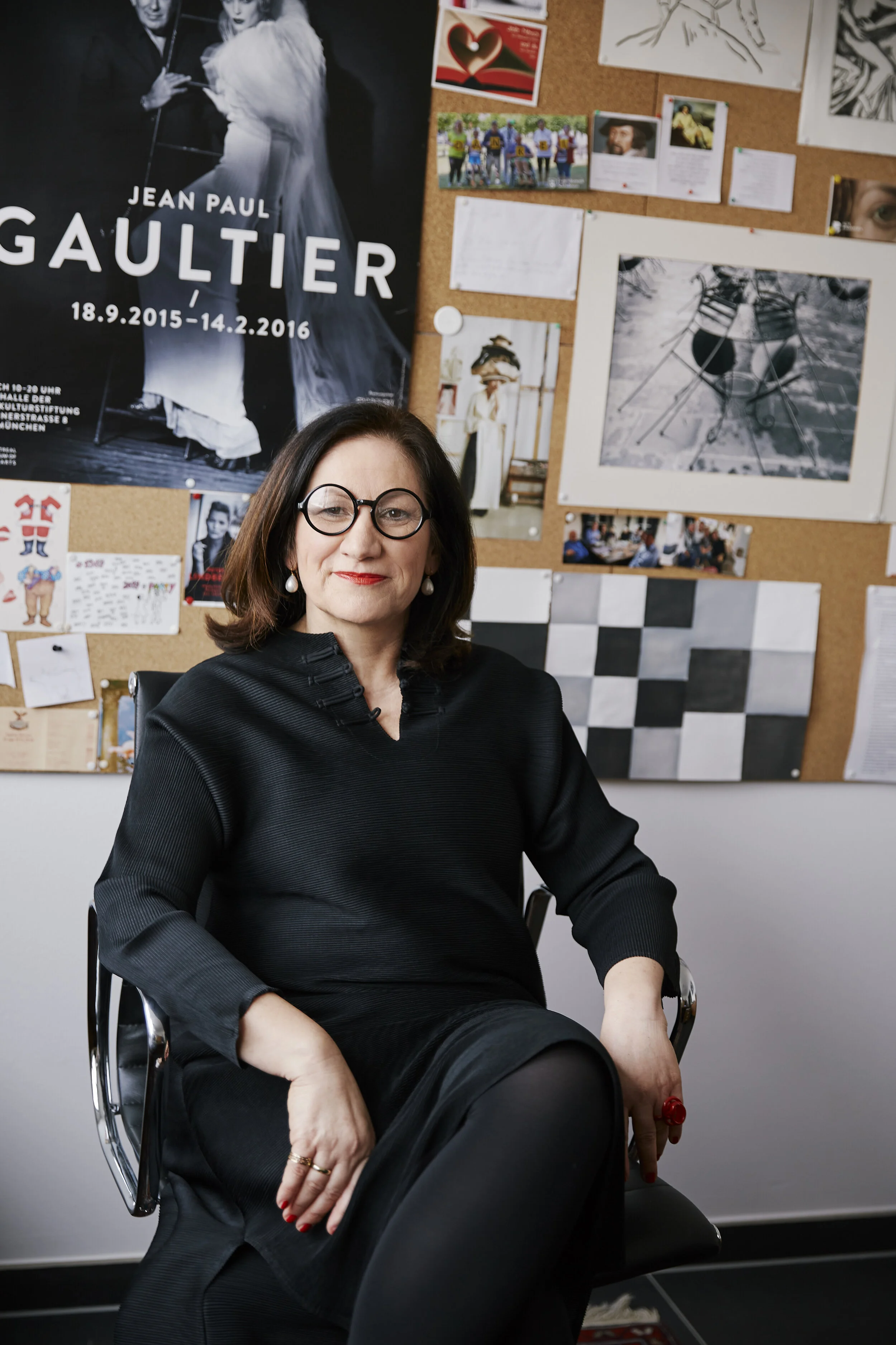 Frau sitzt in einem Büro mit einer Pinnwand im Hintergrund, die mit Bildern und einem großen Jean Paul Gaultier Poster dekoriert ist.