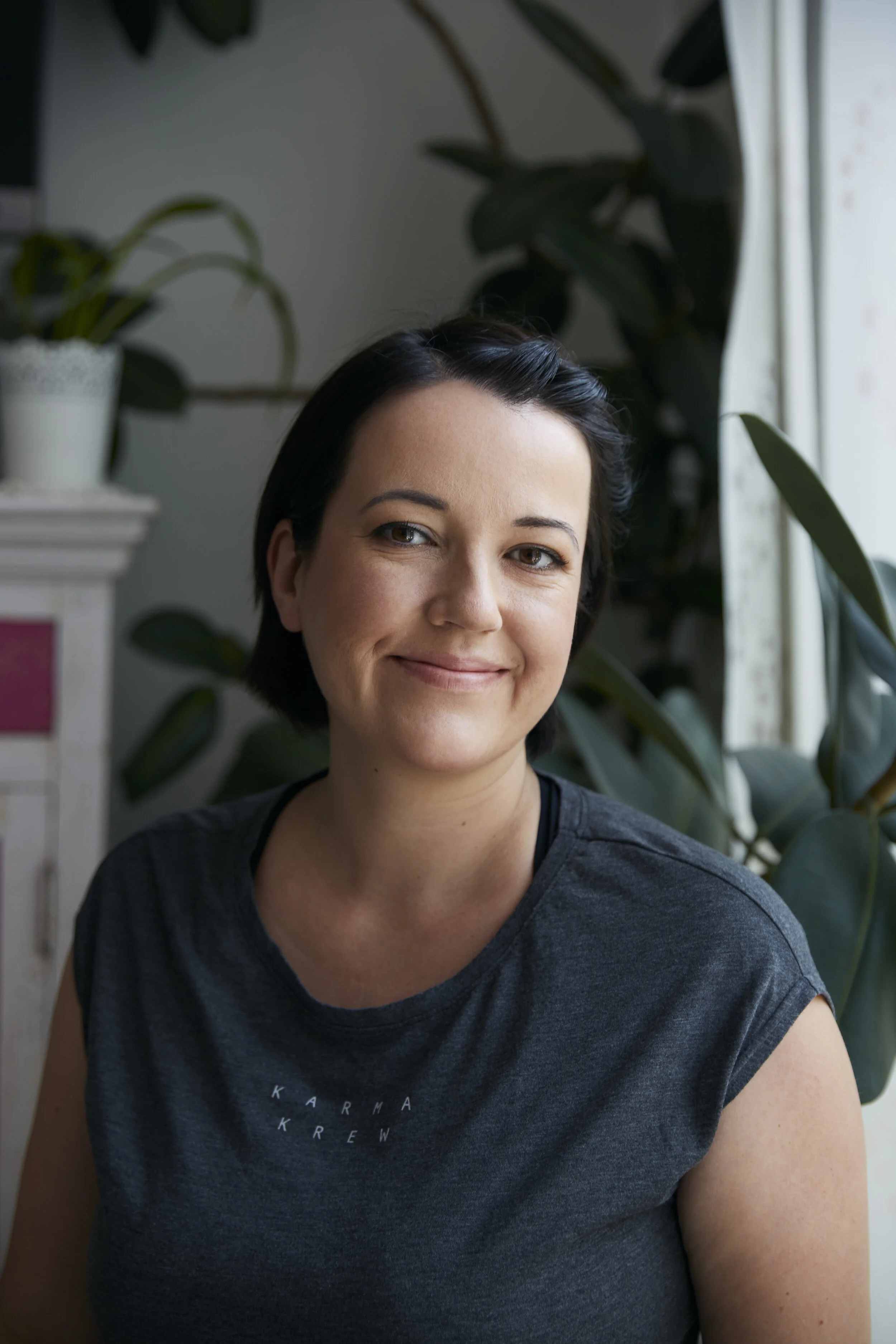 Frau mit kurzem dunklem Haar, lächelt, trägt ein dunkelgraues T-Shirt, Pflanzen im Hintergrund.