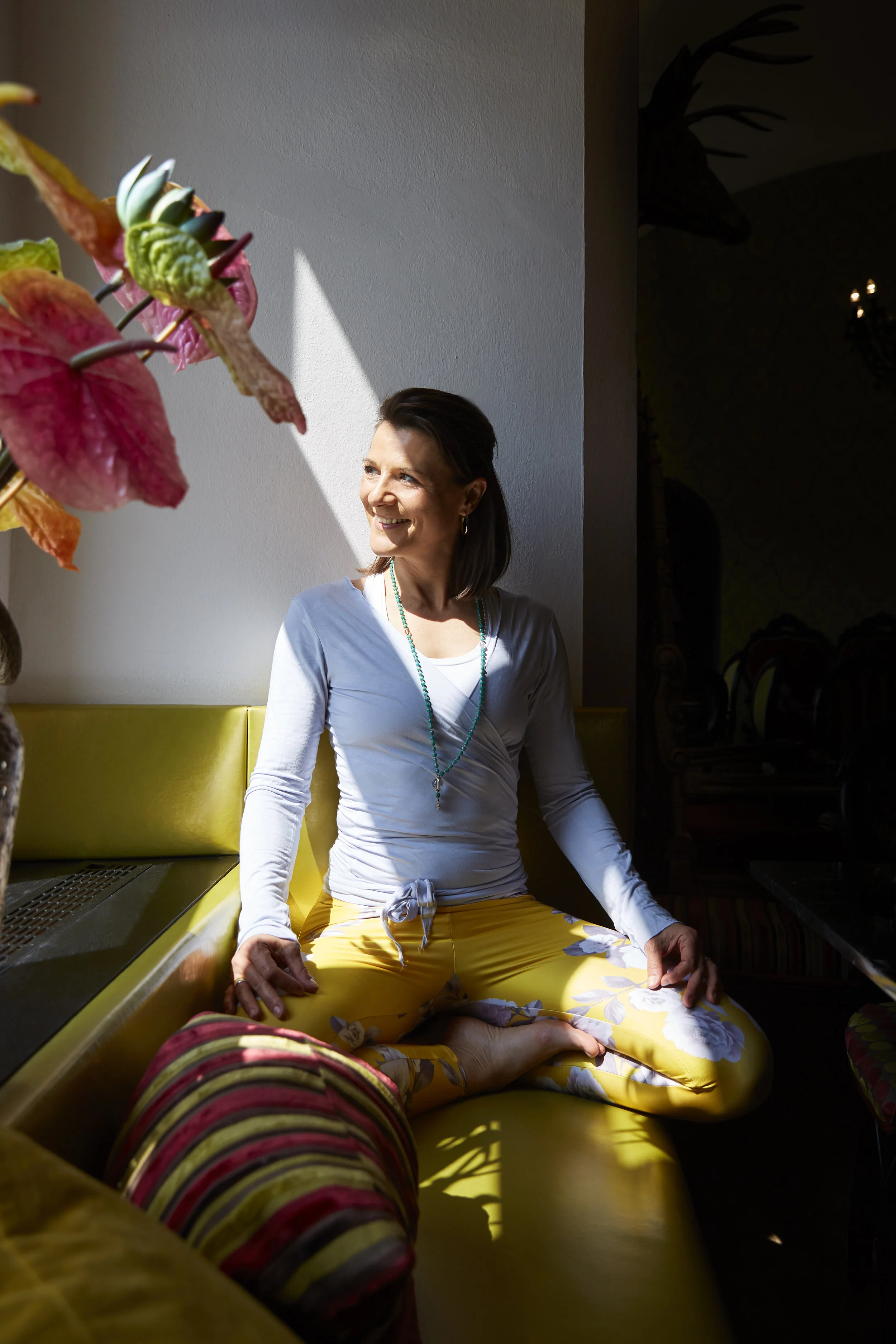 Eine Frau sitzt in einem sonnigen Raum auf einem gelben Sofa. Sie trägt ein weißes Oberteil und gelbe Blumenleggings und schaut lächelnd nach draußen. Im Vordergrund sind bunte Blätter einer Pflanze zu sehen.