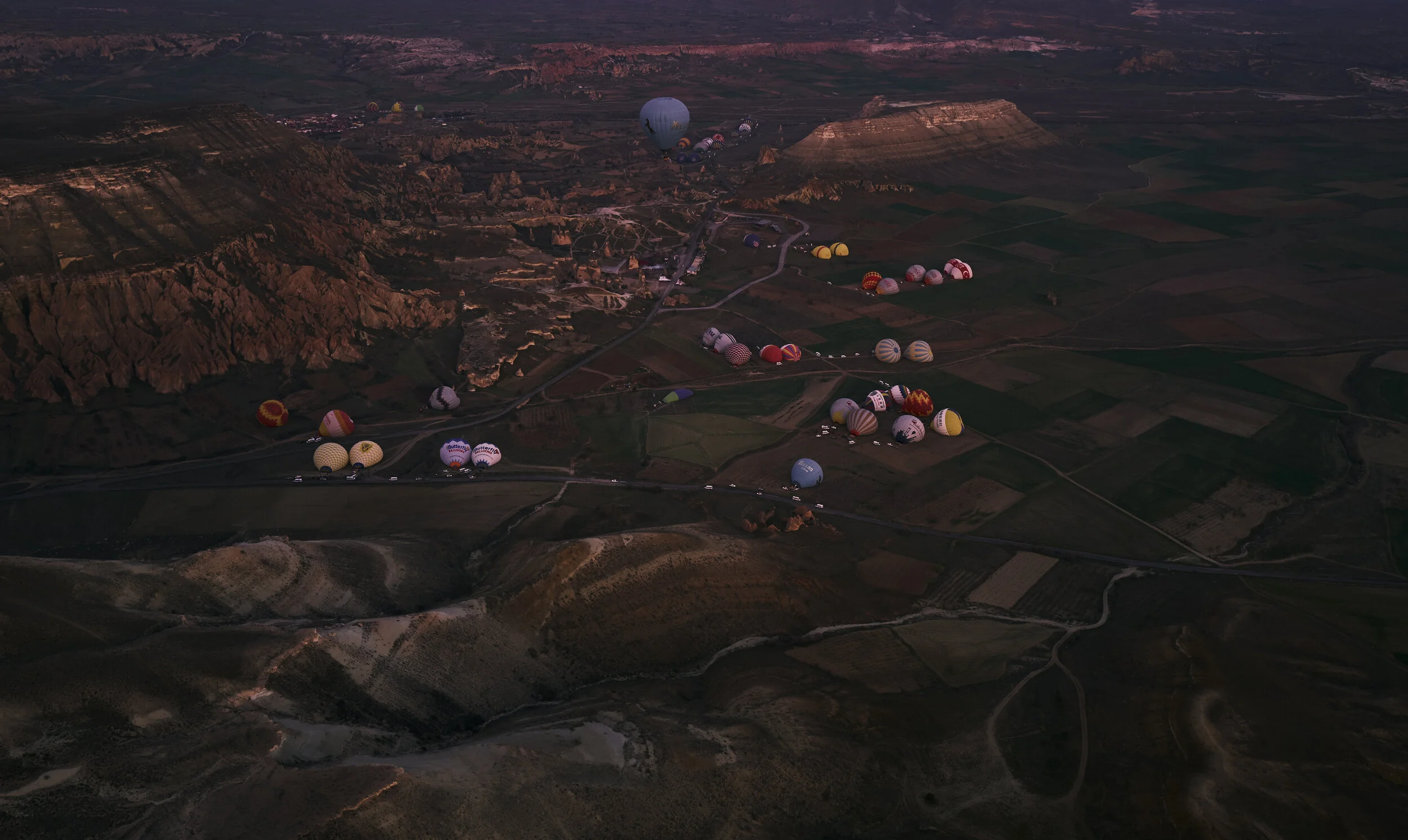 Landschaftsaufnahme mit vielen bunten Heißluftballons, die über ein hügeliges Tal starten. Im Hintergrund sind Felsen und weite Felder sichtbar.