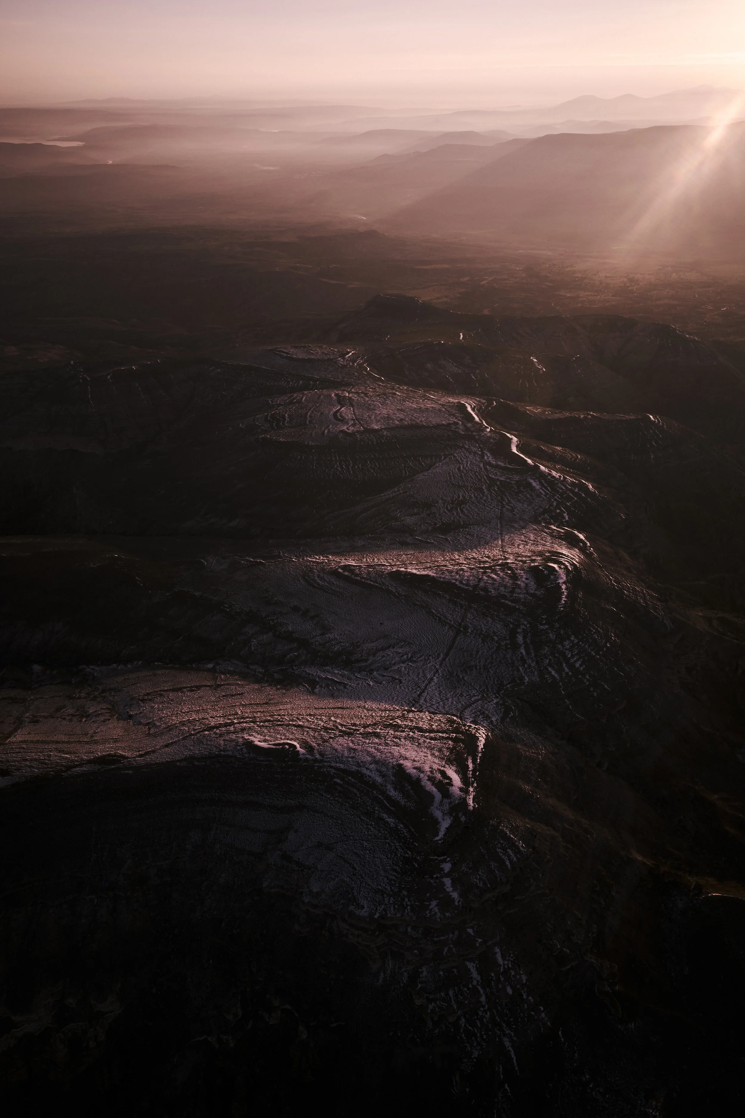 Luftaufnahme einer gebirgigen Landschaft bei Sonnenaufgang mit dunstigem Himmel und Lichtstrahlen auf den Hügeln.