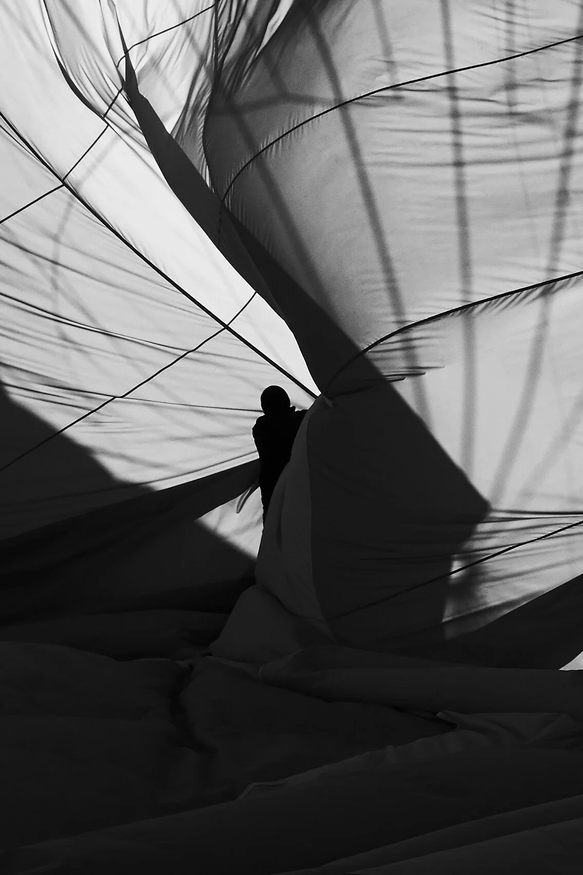 Schwarz-weiß-Foto einer Silhouette einer Person, die in einer großen Stoffbahn steht, möglicherweise in einem aufblasbaren Ballon oder Zelt. Licht und Schatten werfen Muster auf den Stoff.