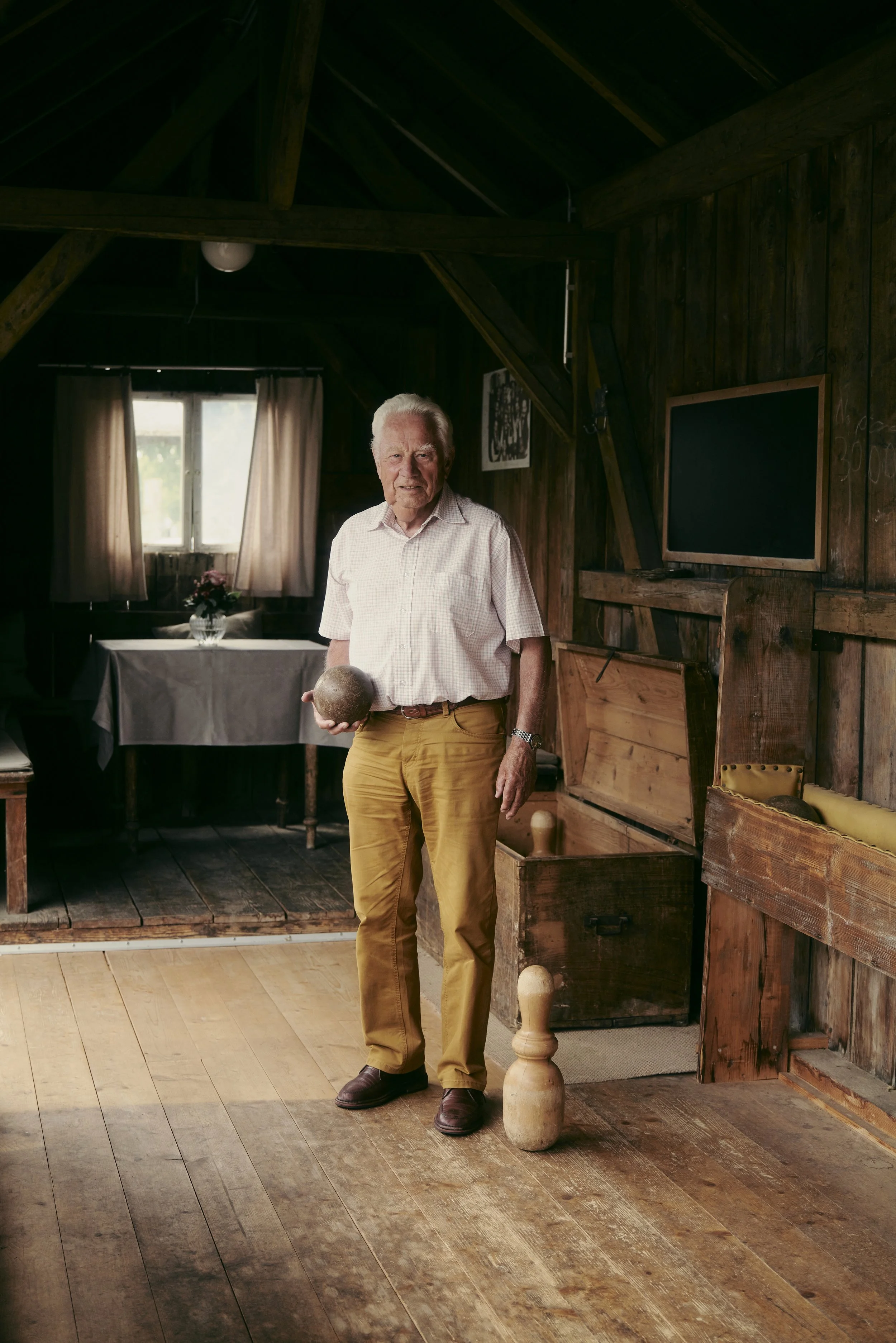Ein älterer Mann steht in einem rustikalen Holzraum mit einer hölzernen Kegelbahn. Er hält eine Kugel in der Hand. Im Hintergrund ist ein Tisch mit einer Blume im Fensterlicht zu sehen.