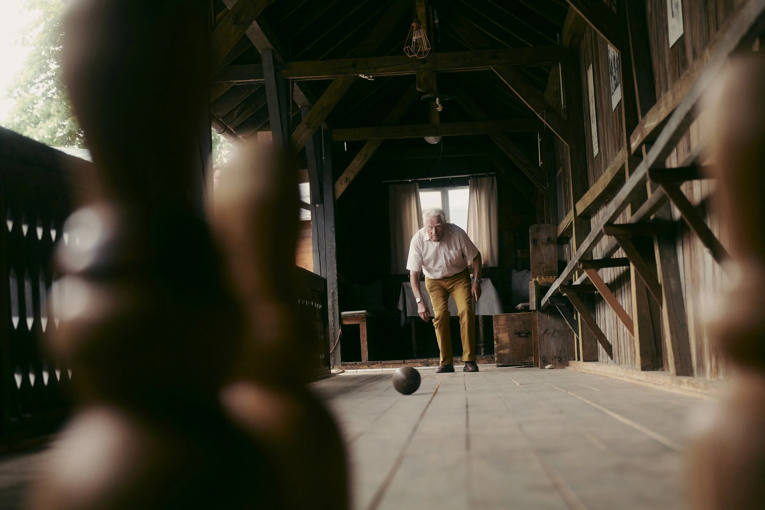 Ein älterer Mann spielt Kegeln auf einer traditionellen Kegelbahn aus Holz, während er eine Kugel wirft. Der Raum ist rustikal und aus Holz gebaut, mit Holzbalken an der Decke.
