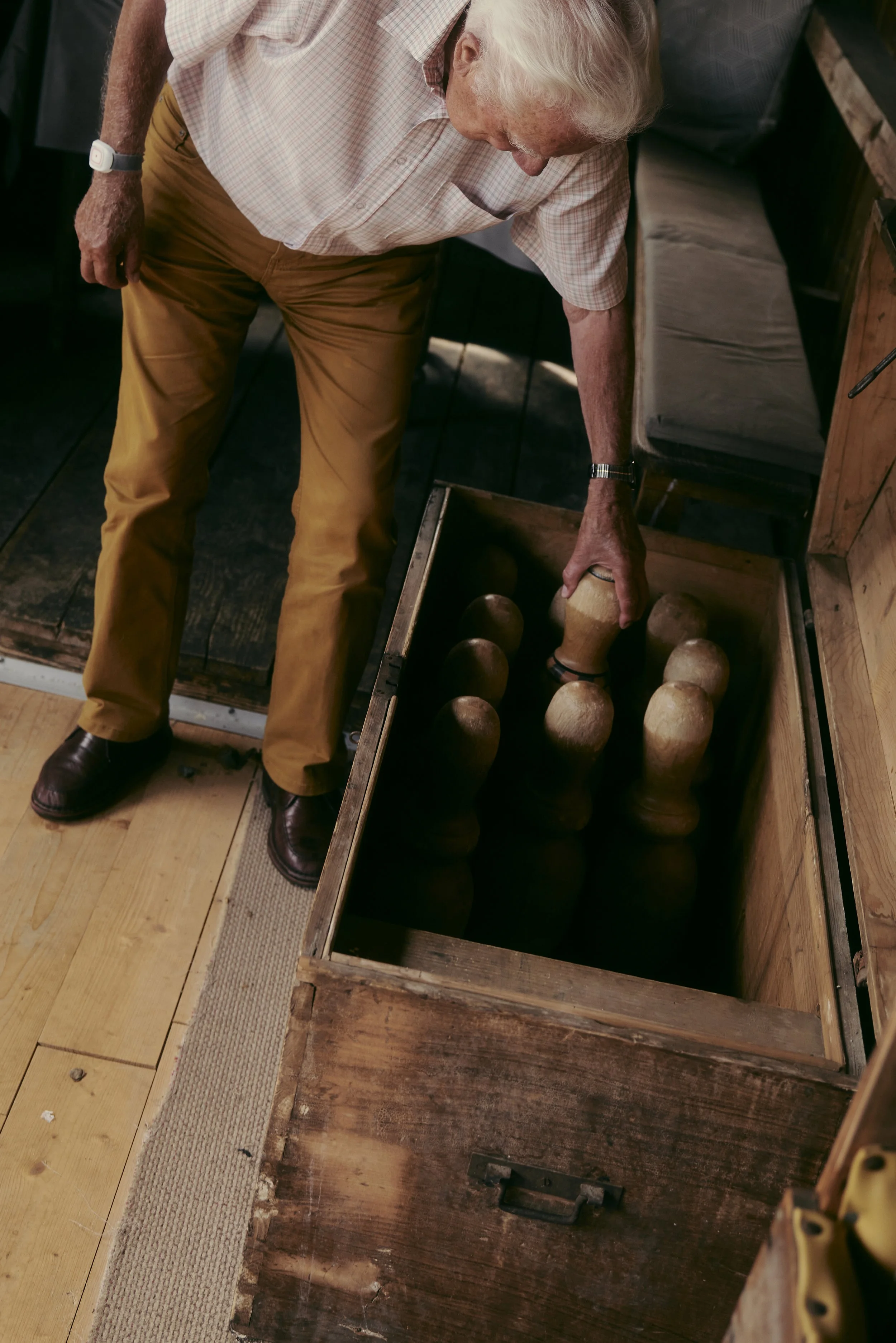 Ein älterer Mann hält eine Holzkegel in einer Holzkiste mit mehreren Kegeln. Er trägt beigen Hosen und ein weiß kariertes Hemd.