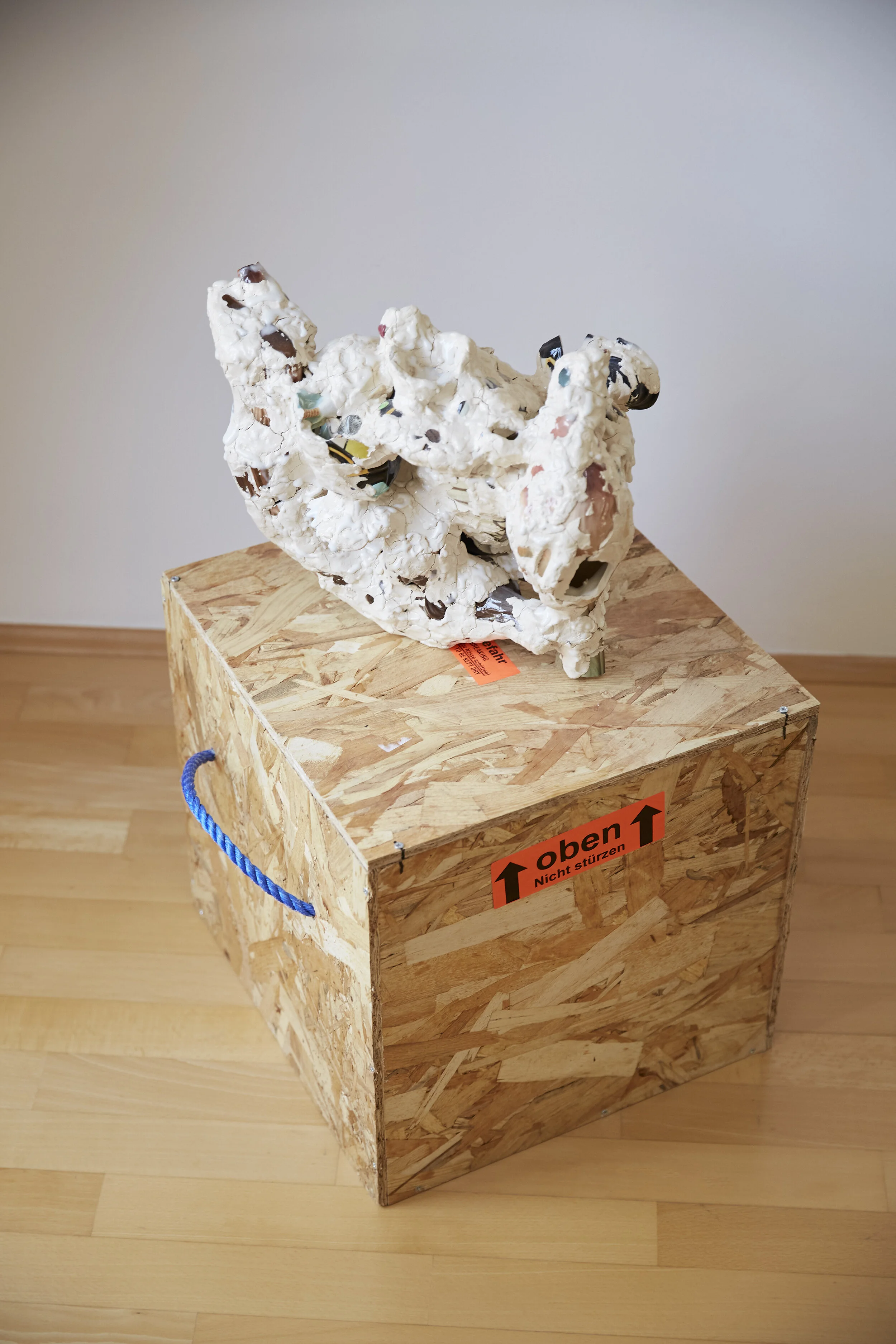 Kunstskulptur aus weißem Material mit Farbsprenkeln auf einem Holzkasten mit Aufschrift 'oben, nicht stürzen'.