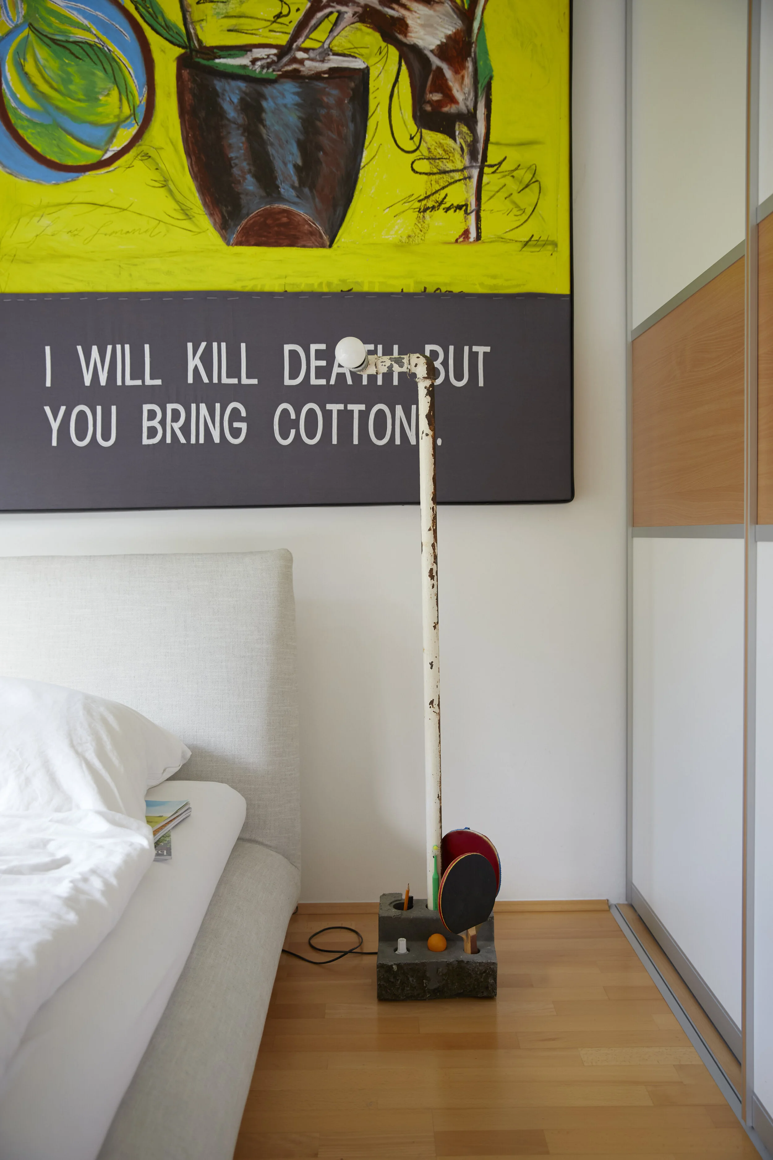 Ein minimalistisches Schlafzimmer mit einem weißen Bettgestell und Kissen. An der Wand hängt ein kunstvolles Bild mit fortgeschrittenem englischem Text. Auf dem Boden steht eine rustikale Stehlampe auf einem Betonblock, daneben einige Tischtennisschl