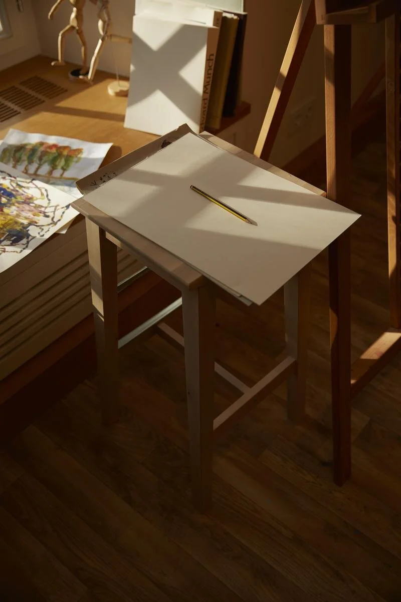 Ein Holzhocker mit einem Blatt Papier und einem Bleistift darauf. Im Hintergrund eine Zeichnung und Bücher an einem Fensterbrett. Holzfußboden und Sonnenlicht.