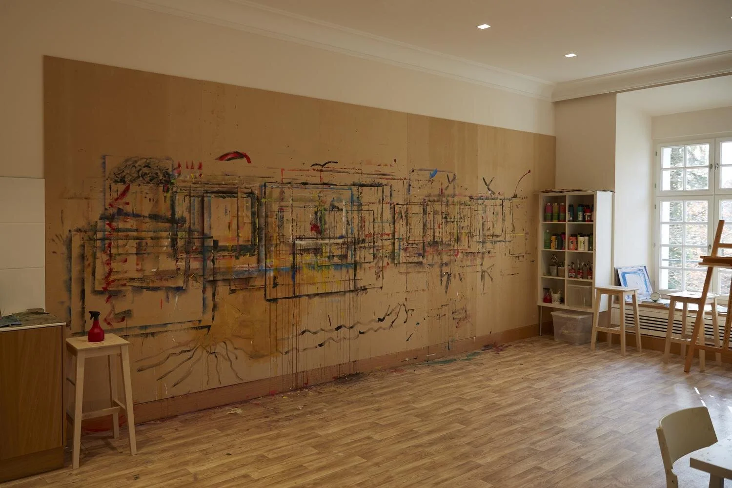 Ein Kunstatelier mit einer großen Leinwand an der Wand, auf der abstrakte Kunst mit verschiedenen Farben, Linien und Formen gemalt ist. Im Raum stehen Hocker, ein Regal mit Malutensilien und ein großes Fenster, das Licht hereinlässt.