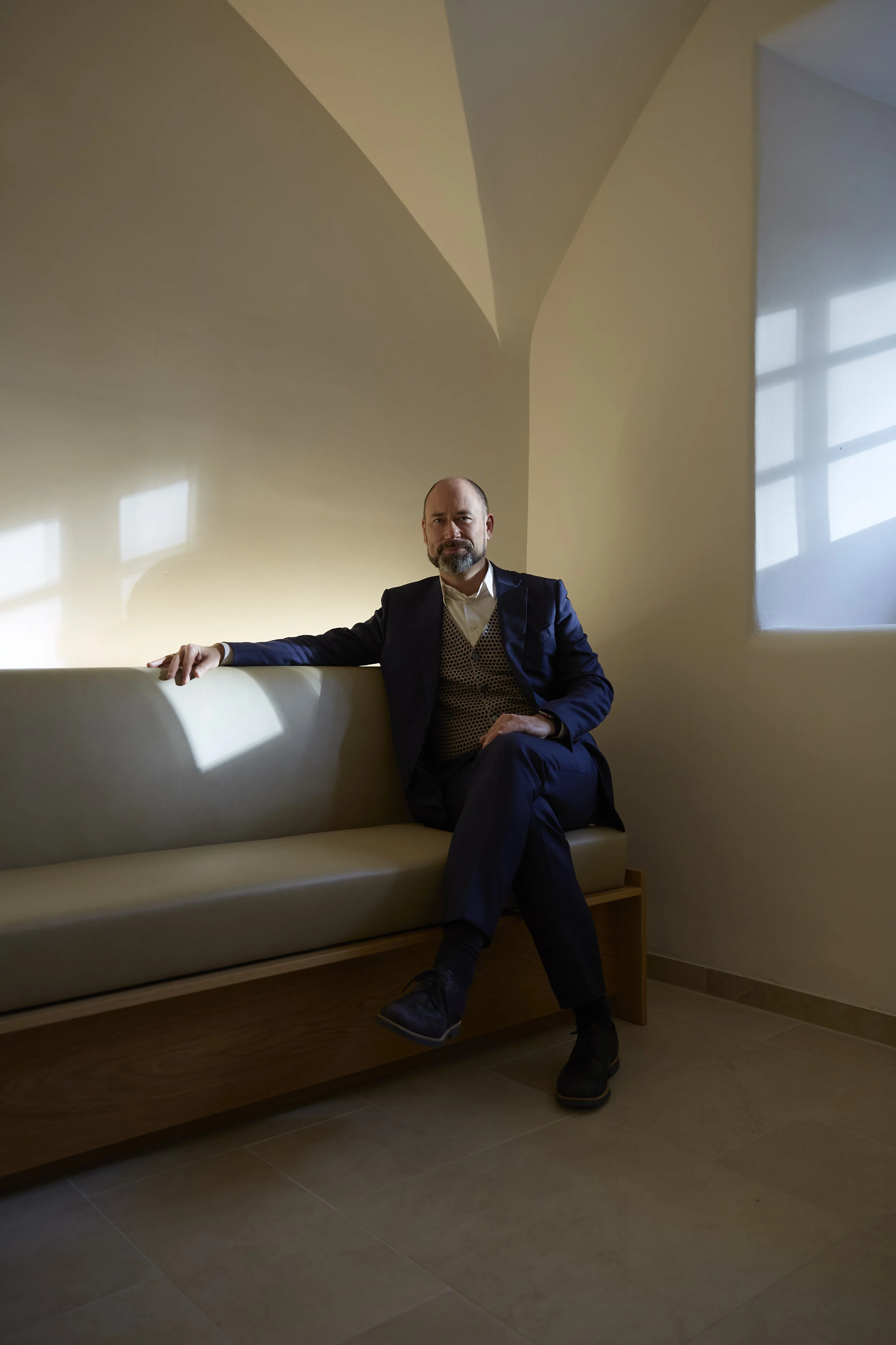 Mann im Anzug sitzt auf einer Bank in einem minimalistischen Raum, beleuchtet von Sonnenlicht durch ein Fenster.