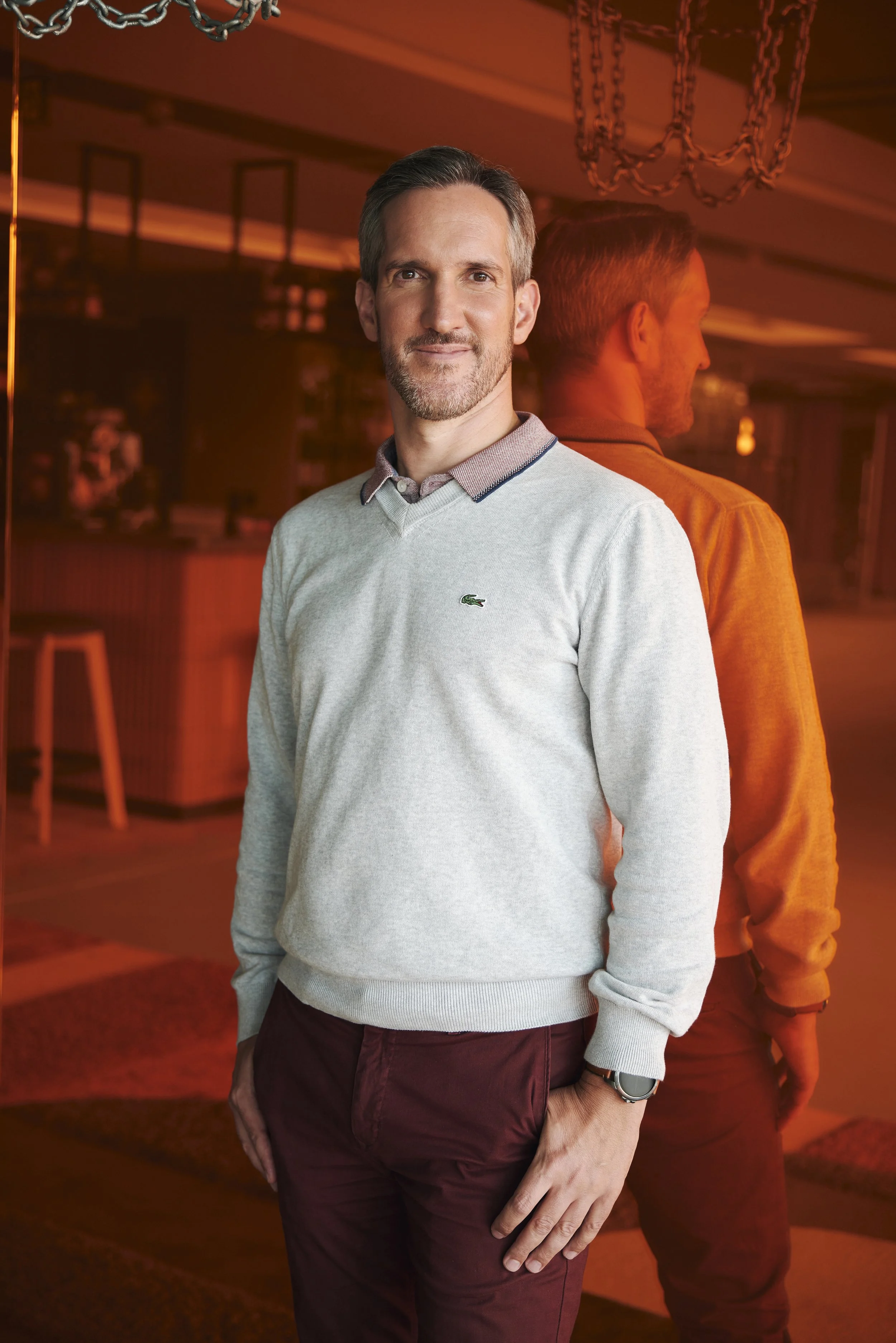 Mann in grauem Pullover und bordeauxroten Hosen in einem Innenraum, hinter ihm ein Spiegel.