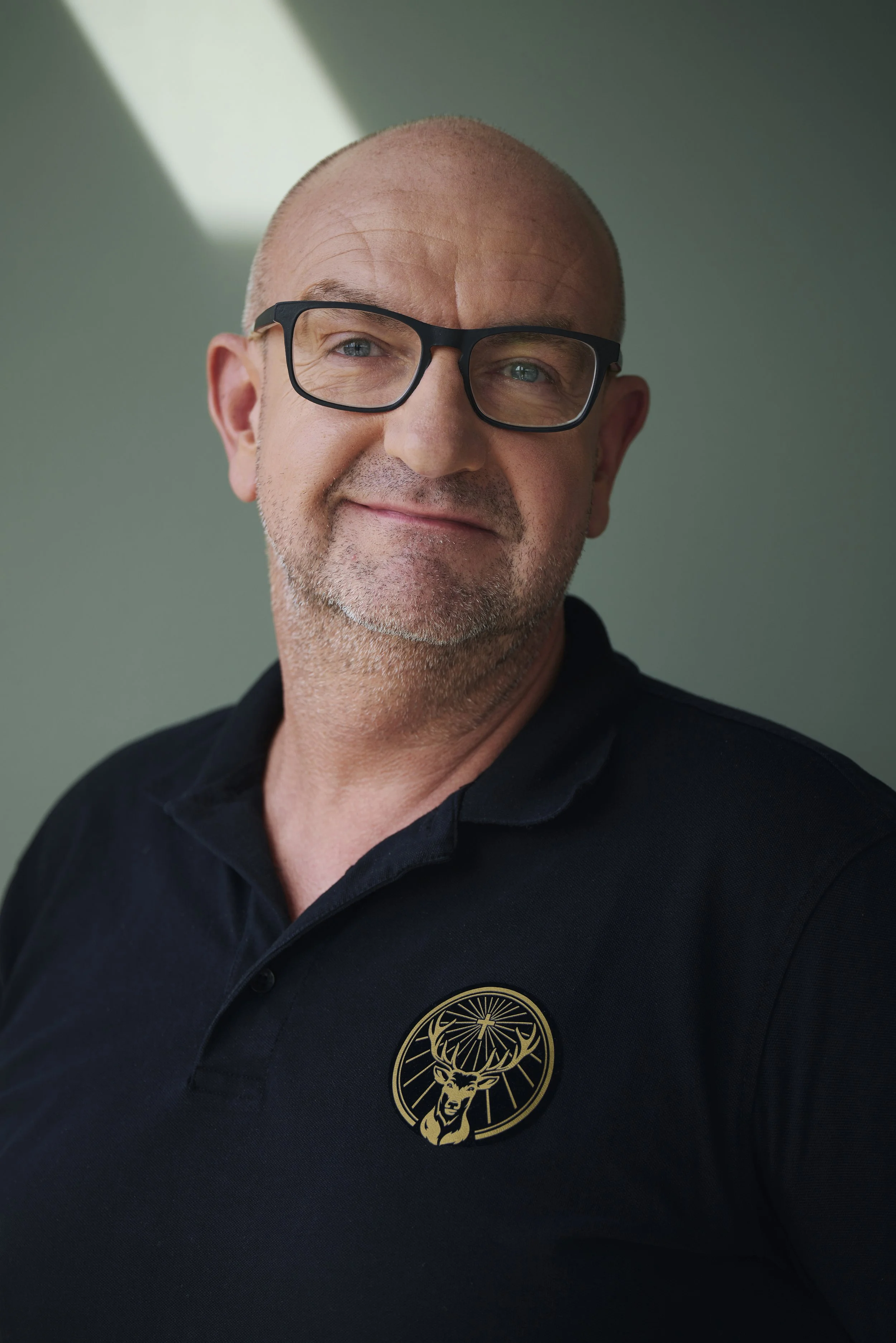 Mann mit Brille und schwarzem Polohemd mit Hirschlogo, Hintergrund grün.