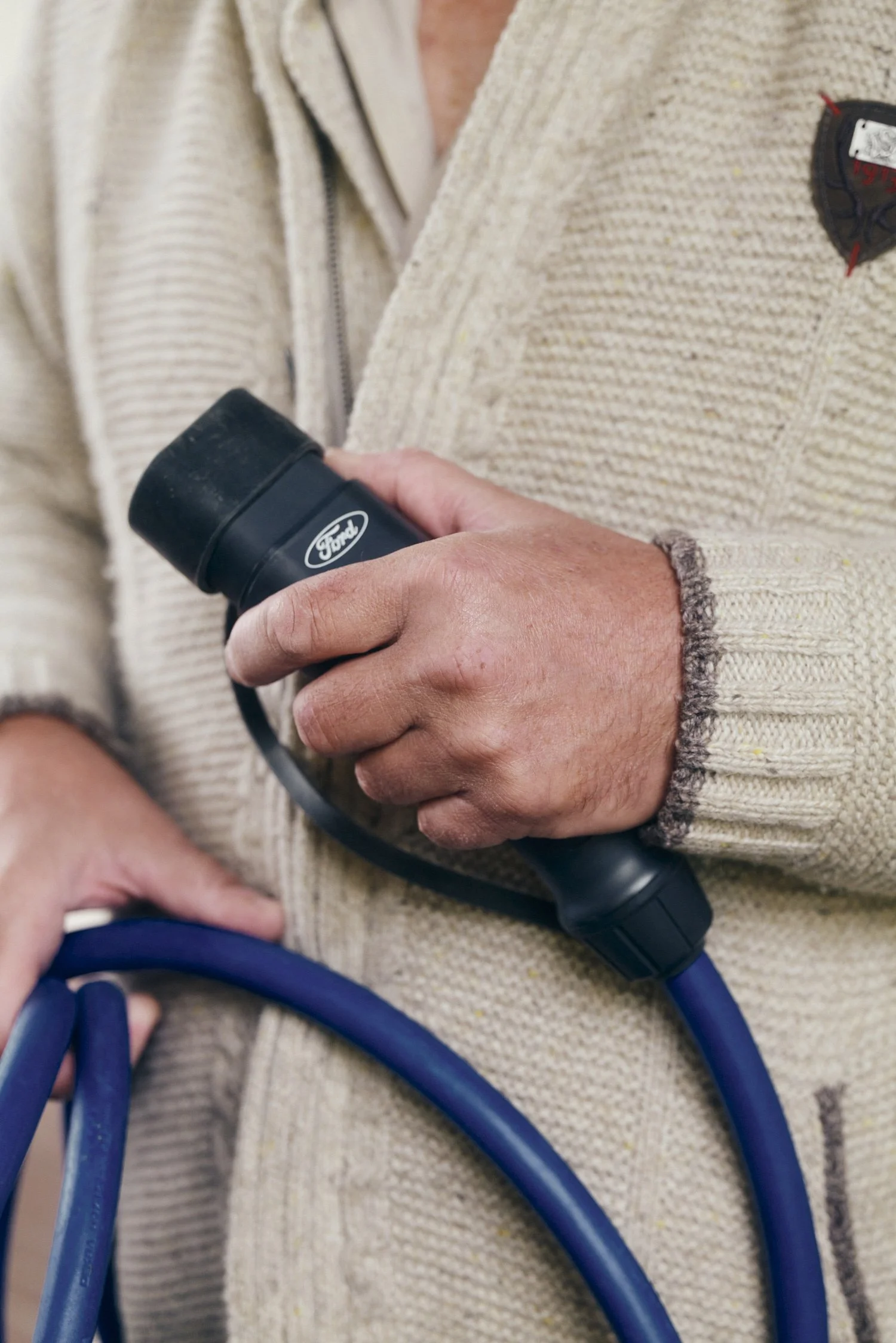 Person in cremefarbenem Strickpullover hält ein blaues Kabel mit einem schwarzen Ford-Stecker.