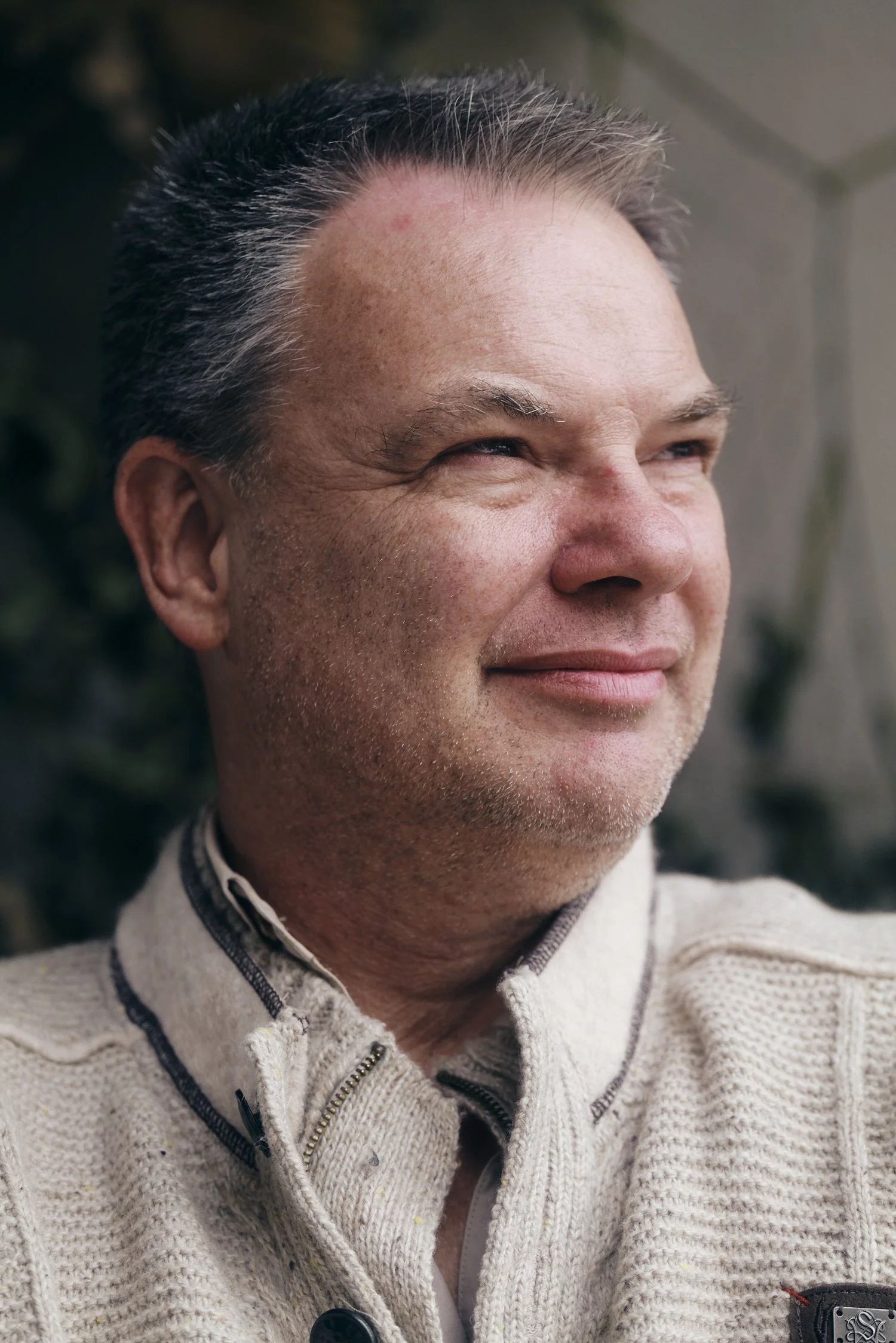 Porträt eines lächelnden Mannes in einem beigen Strickpullover, der nach rechts schaut.