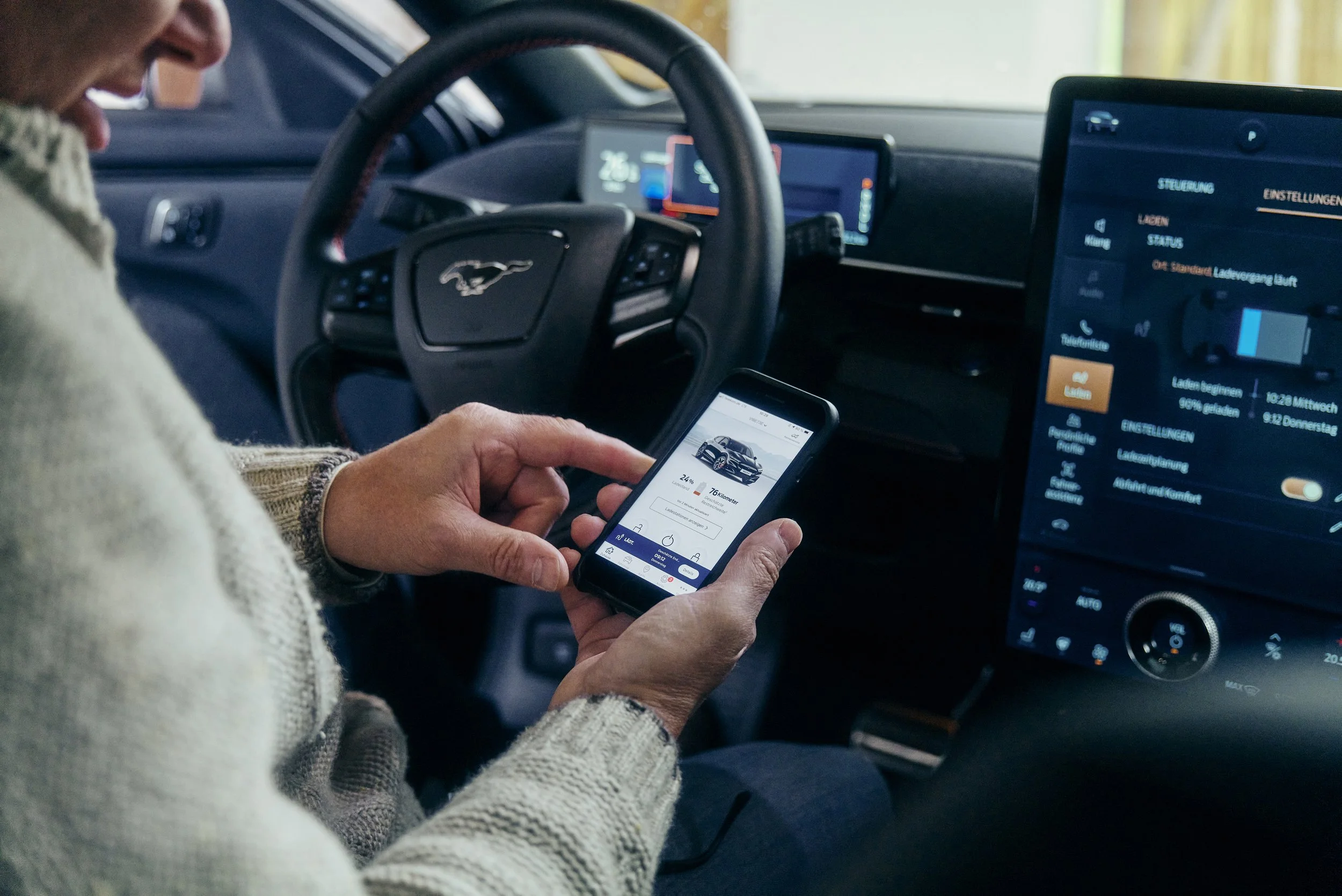 Eine Person sitzt in einem Auto und bedient ein Smartphone. Auf dem Armaturenbrett sind ein Lenkrad mit einem Mustang-Logo und ein großes Display mit Menüoptionen sichtbar.