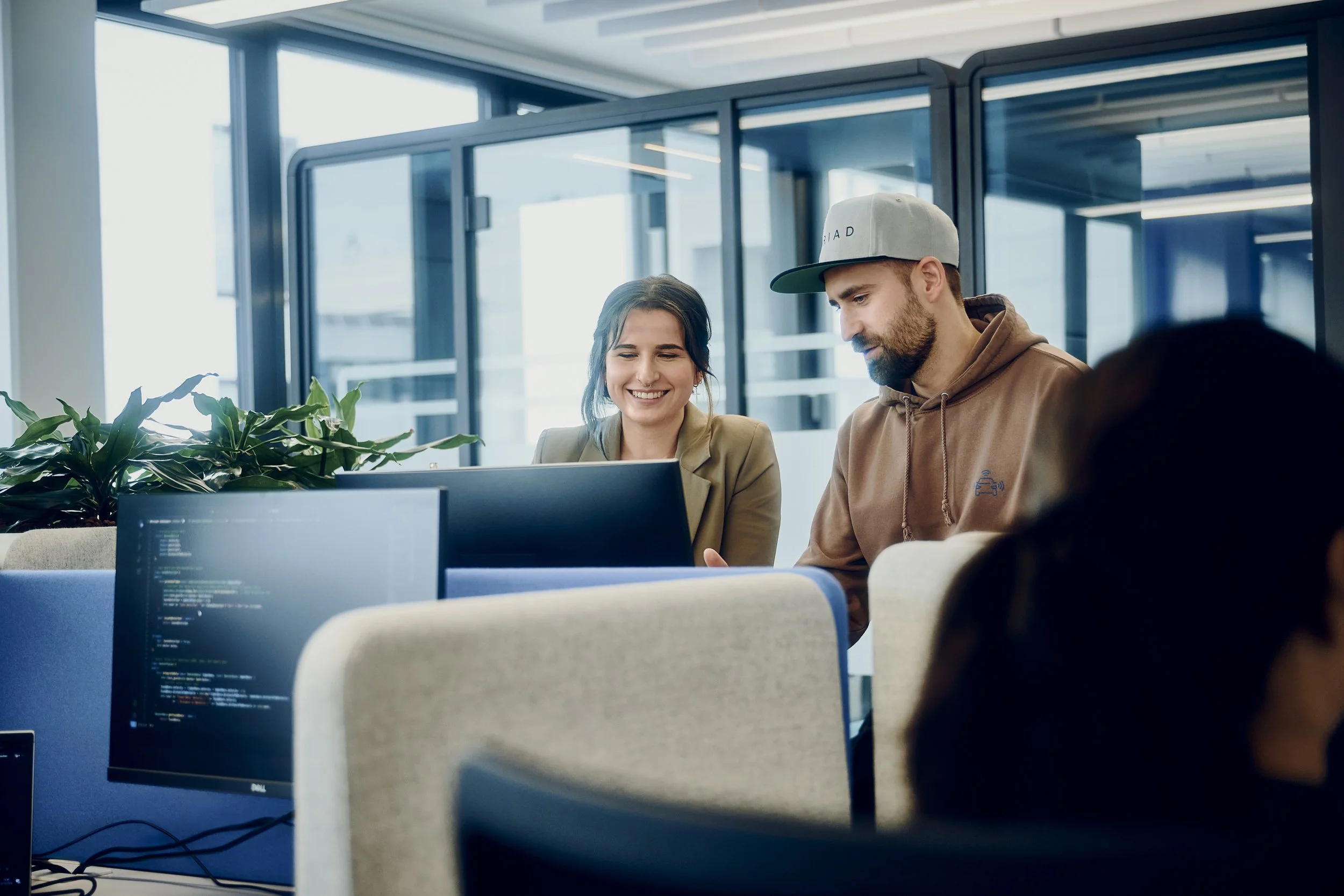 Zwei Personen arbeiten zusammen an einem Computer in einem modernen Büro. Sie lächeln und scheinen sich auf den Bildschirm zu konzentrieren. Im Vordergrund sind Codezeilen auf einem Monitor zu sehen. Pflanzen und eine gläserne Trennwand sind im Hintergrund.