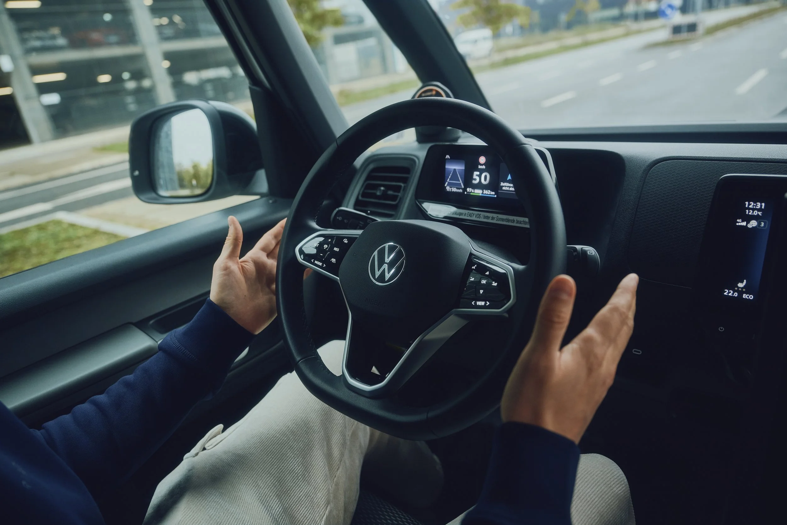 Eine Person sitzt in einem Auto und hält das Lenkrad, das ein Volkswagen-Logo zeigt. Der Armaturenbrettbildschirm zeigt Fahrinformationen. Der Außenspiegel und die Straße sind durch die Seitenscheibe sichtbar. Die Hände der Person sind nahe am Lenkrad, während sie vorausschauend fährt.