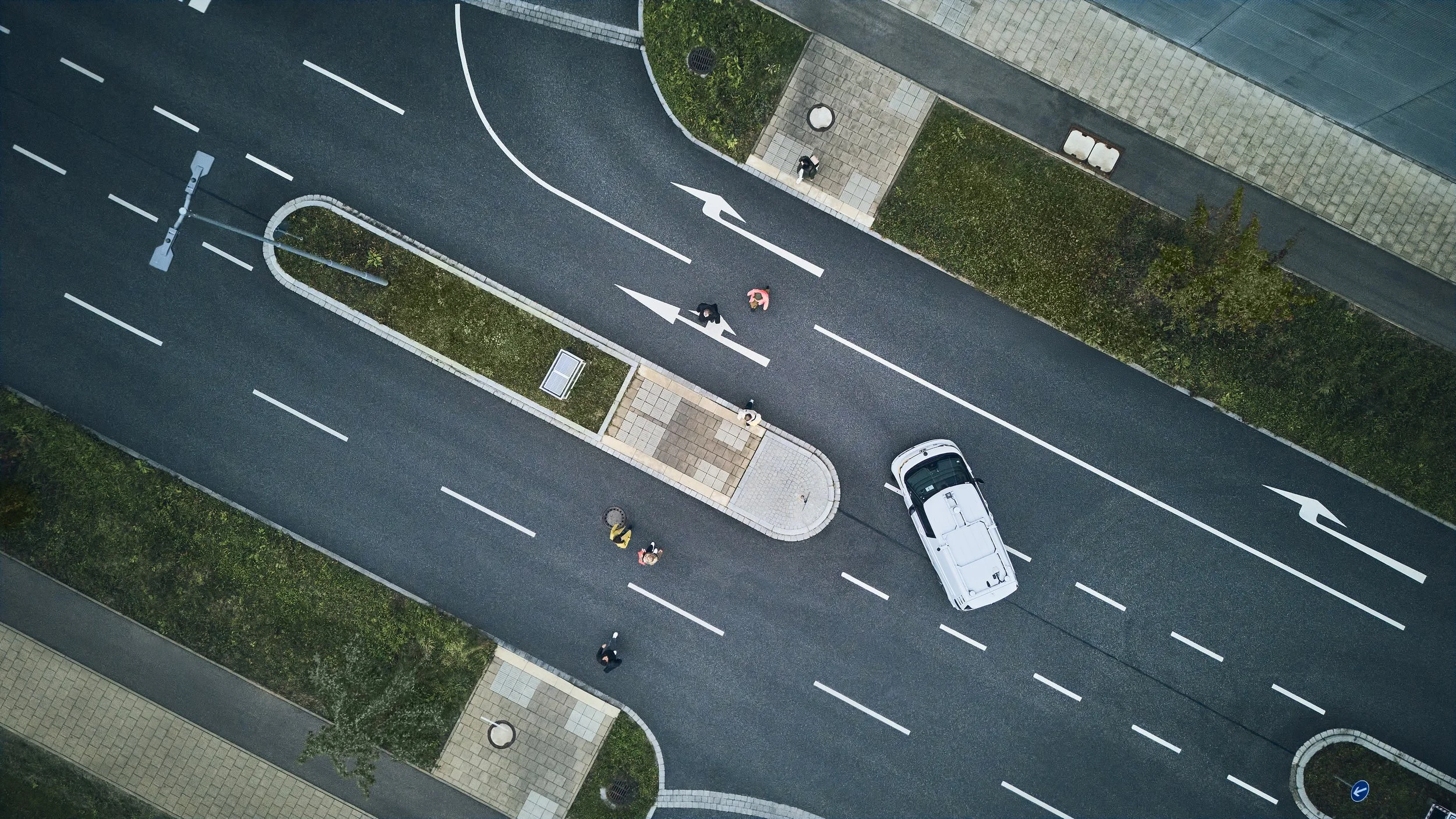 Luftaufnahme einer Kreuzung mit einem weißen Auto und mehreren Fußgängern auf der Straße und Gehwegen. Straßenmarkierungen sind sichtbar, darunter Pfeile, die die Fahrtrichtung anzeigen. Grünflächen sind entlang der Straßen.