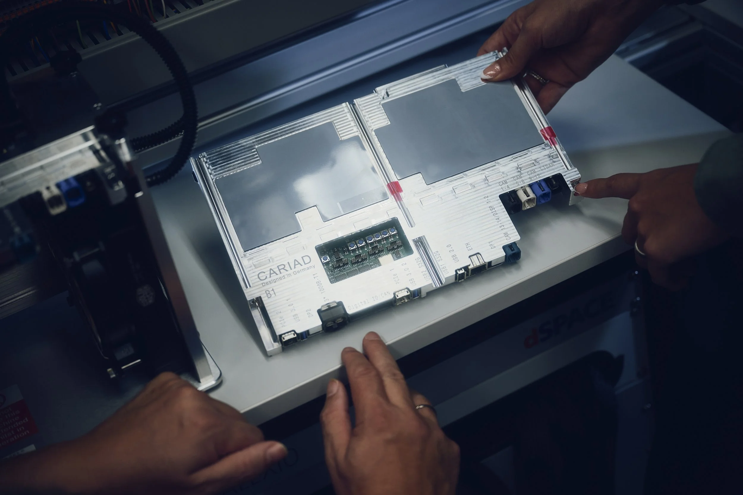 Personen halten ein technisches Gerät mit der Aufschrift "CARIAD" auf einem Tisch. Der Raum wirkt wie ein Labor oder Werkstatt.