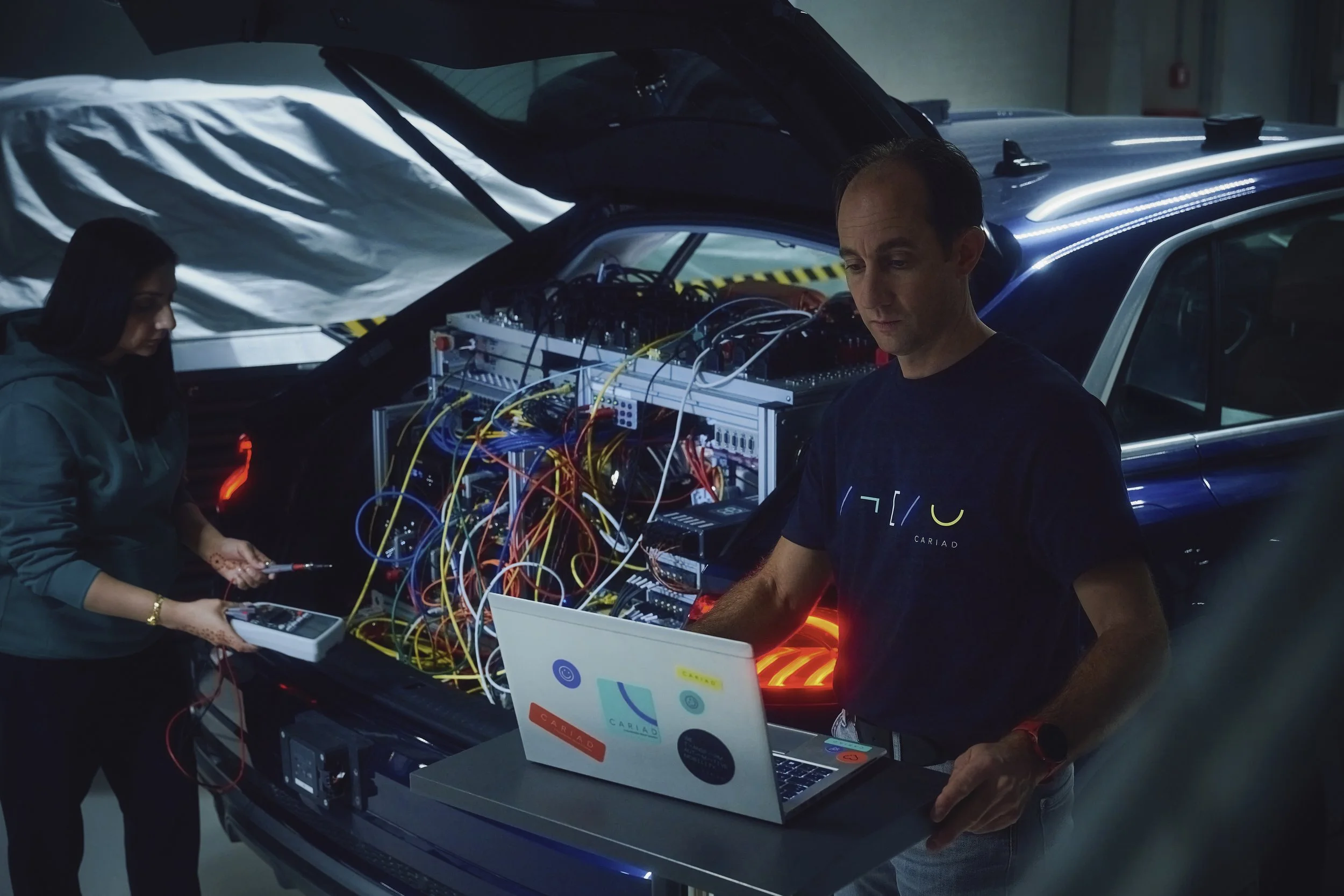 Zwei Personen arbeiten an einem Fahrzeug, das mit Computerausrüstung und vielen Kabeln im Kofferraum ausgestattet ist. Eine Person nutzt einen Laptop, während die andere Geräte überprüft.