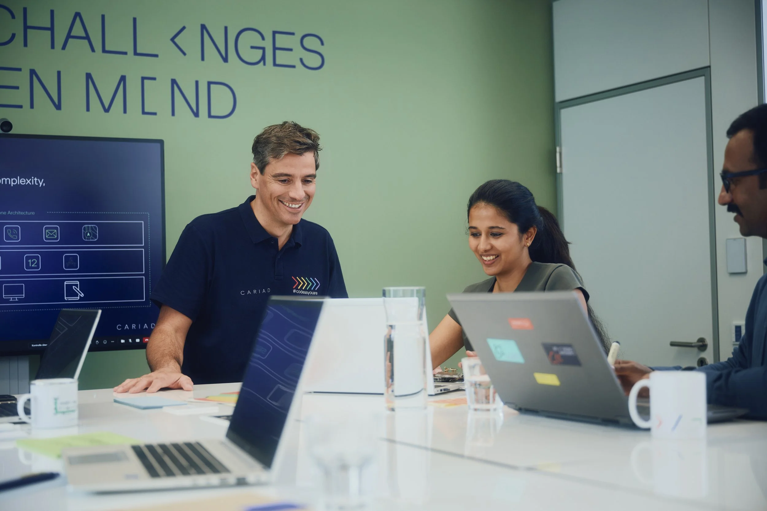 Menschen arbeiten gemeinsam an einem Tisch mit Laptops und einem Bildschirm im Hintergrund. Sie scheinen an einer Besprechung oder einem Teamprojekt teilzunehmen. Der Raum hat eine grüne Wand mit Schriftzug. Alle sind lächelnd konzentriert.