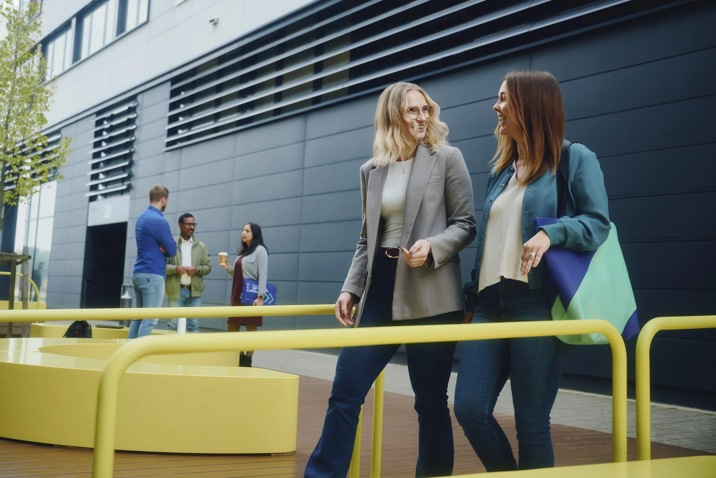 Menschen unterhalten sich draußen vor einem modernen Bürogebäude. Zwei Frauen gehen lächelnd im Vordergrund, während sich im Hintergrund ein Mann und zwei Frauen unterhalten. Gelbe Sitzgelegenheiten und gräuliche Gebäudefassade.