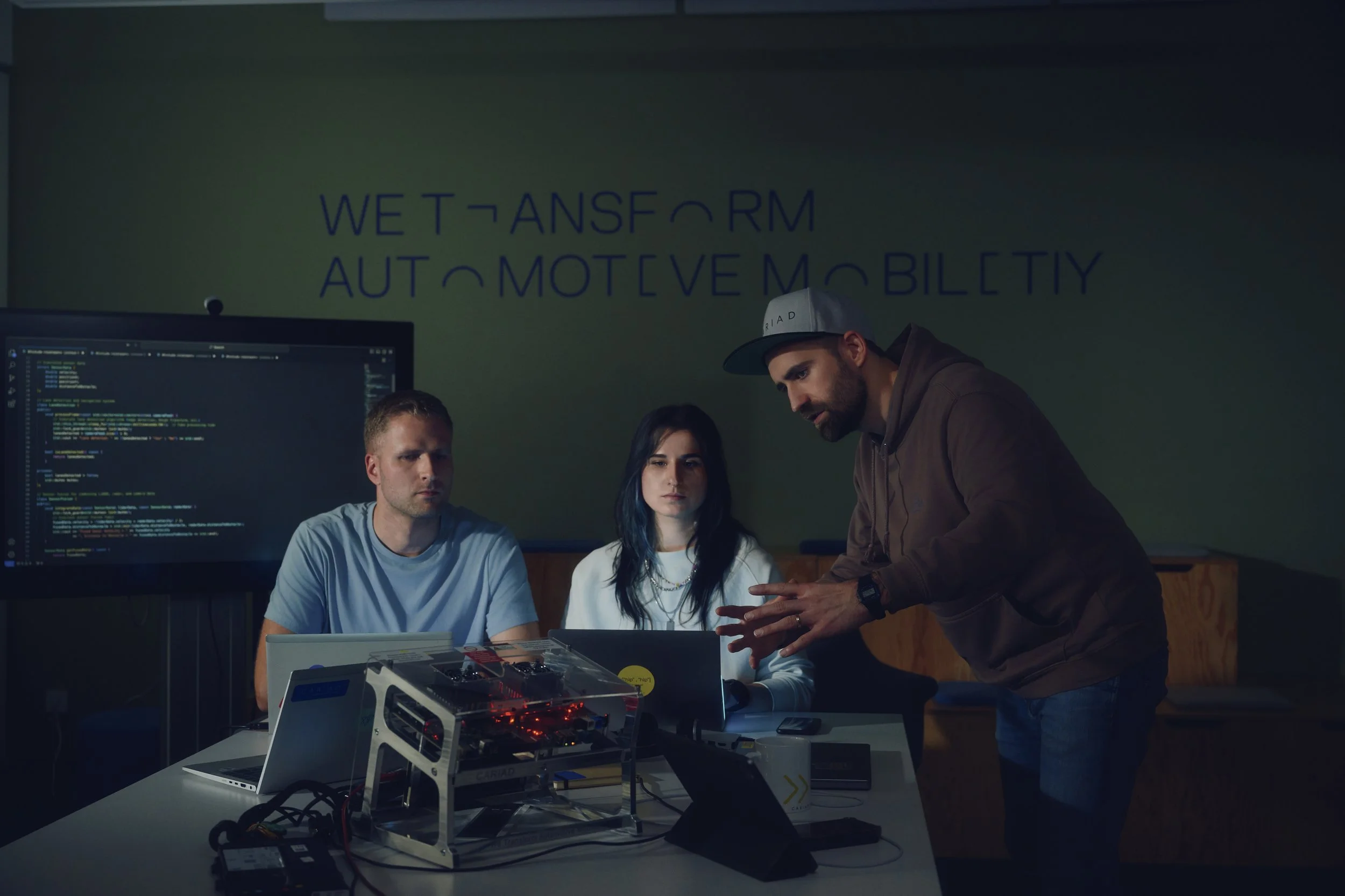 Drei Personen in einem modernen Büro mit Laptops und Technik-Setup, arbeiten an einem Projekt. Im Hintergrund ist ein Bildschirm mit Code zu sehen und ein Schriftzug "We Transform Automotive Mobility" an der Wand.