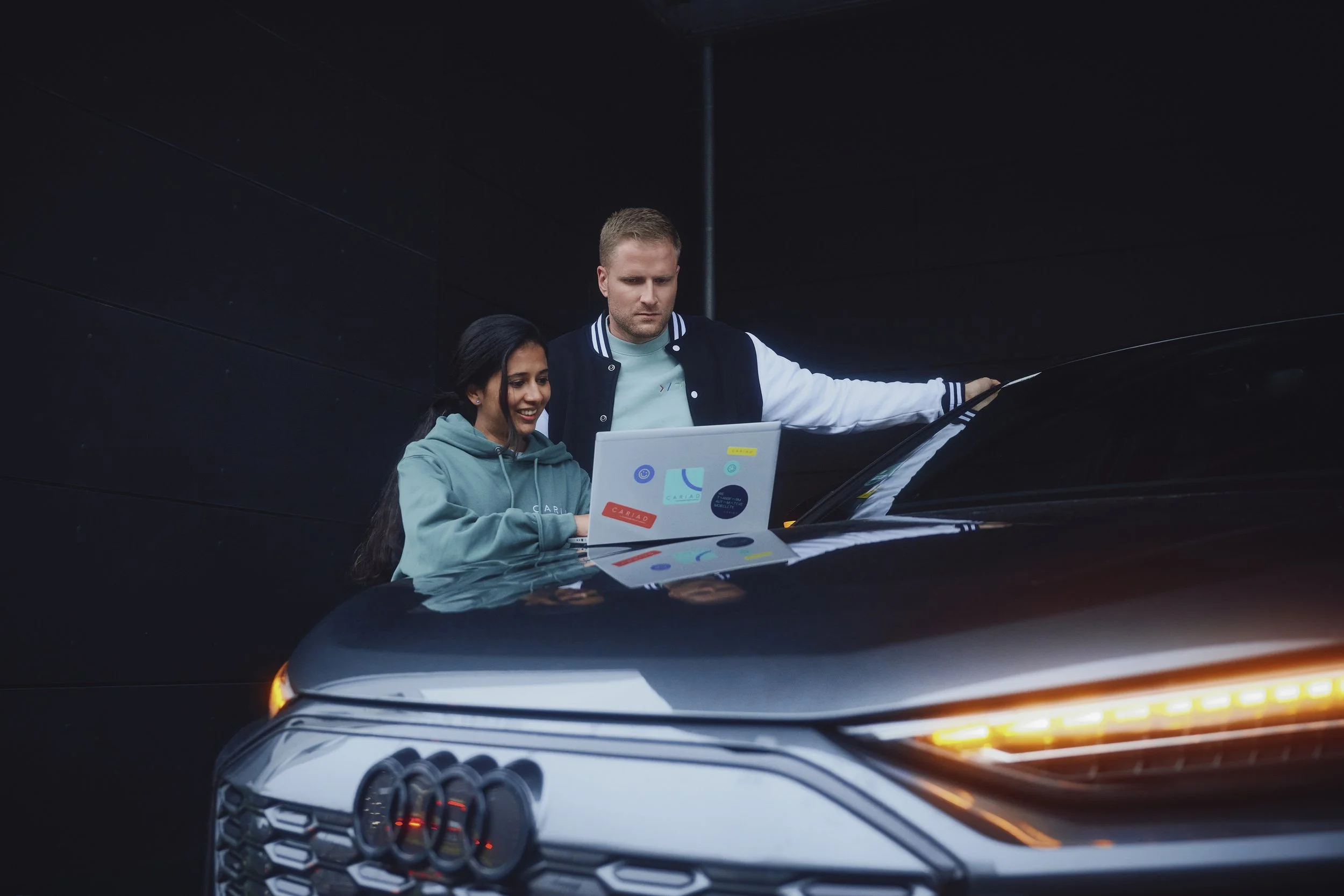 Ein Mann und eine Frau schauen zusammen auf einen Laptop, der auf der Motorhaube eines Audi-Autos liegt. Sie stehen in einem dunklen Raum mit geringer Beleuchtung. Der Mann trägt eine Collegejacke, die Frau einen Kapuzenpullover. Der Laptop ist mit bunten Aufklebern verziert.