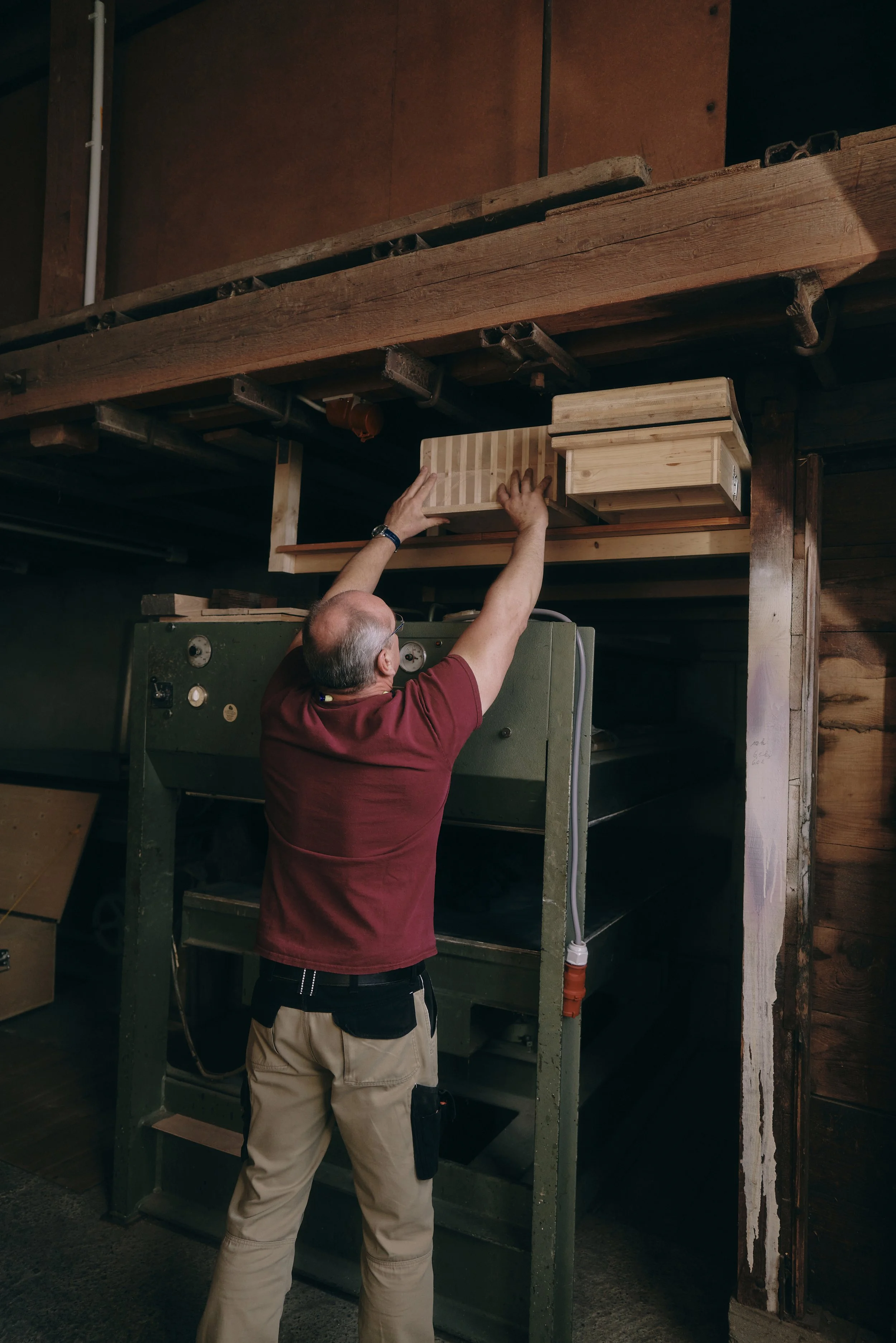 Ein Mann in einem roten T-Shirt und beigen Hosen arbeitet in einer Werkstatt. Er erreicht mit seinen Händen nach einem hölzernen Regal über ihm. Im Hintergrund ist ein großes grünes Gerät oder eine Maschine zu sehen.