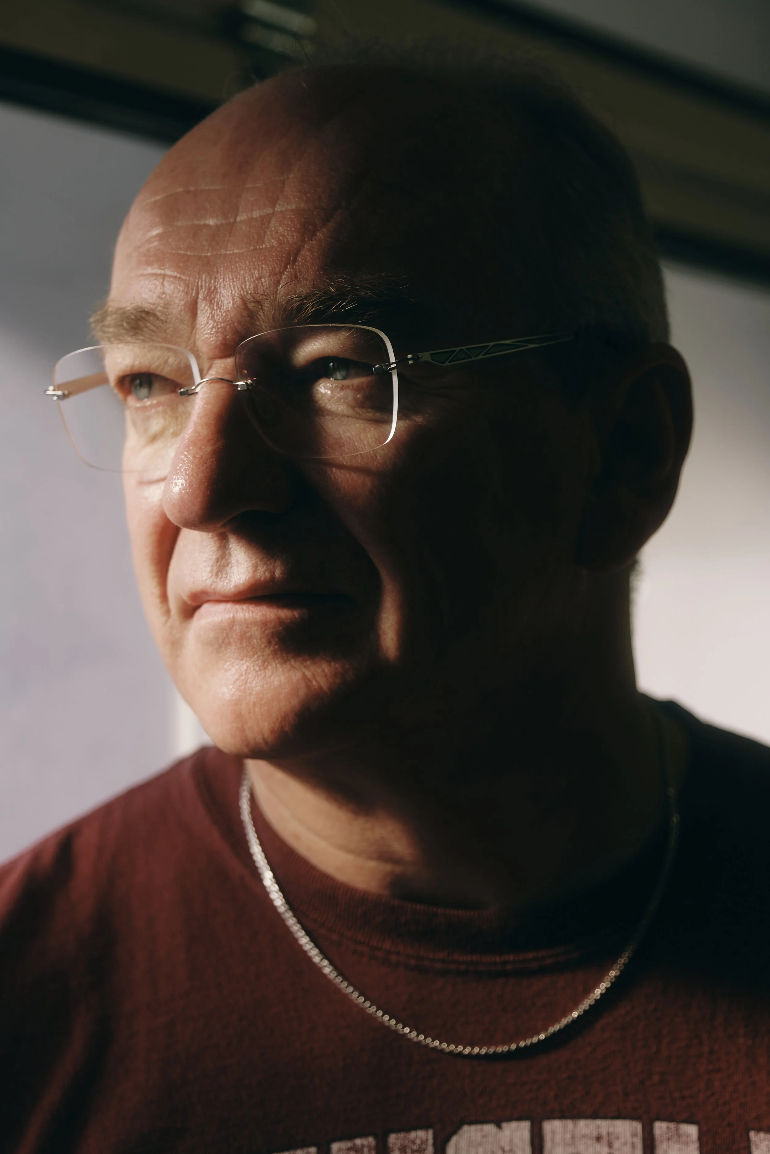 Mann mit Brille und Glatze, trägt ein T-Shirt und eine Kette, schaut nach rechts.