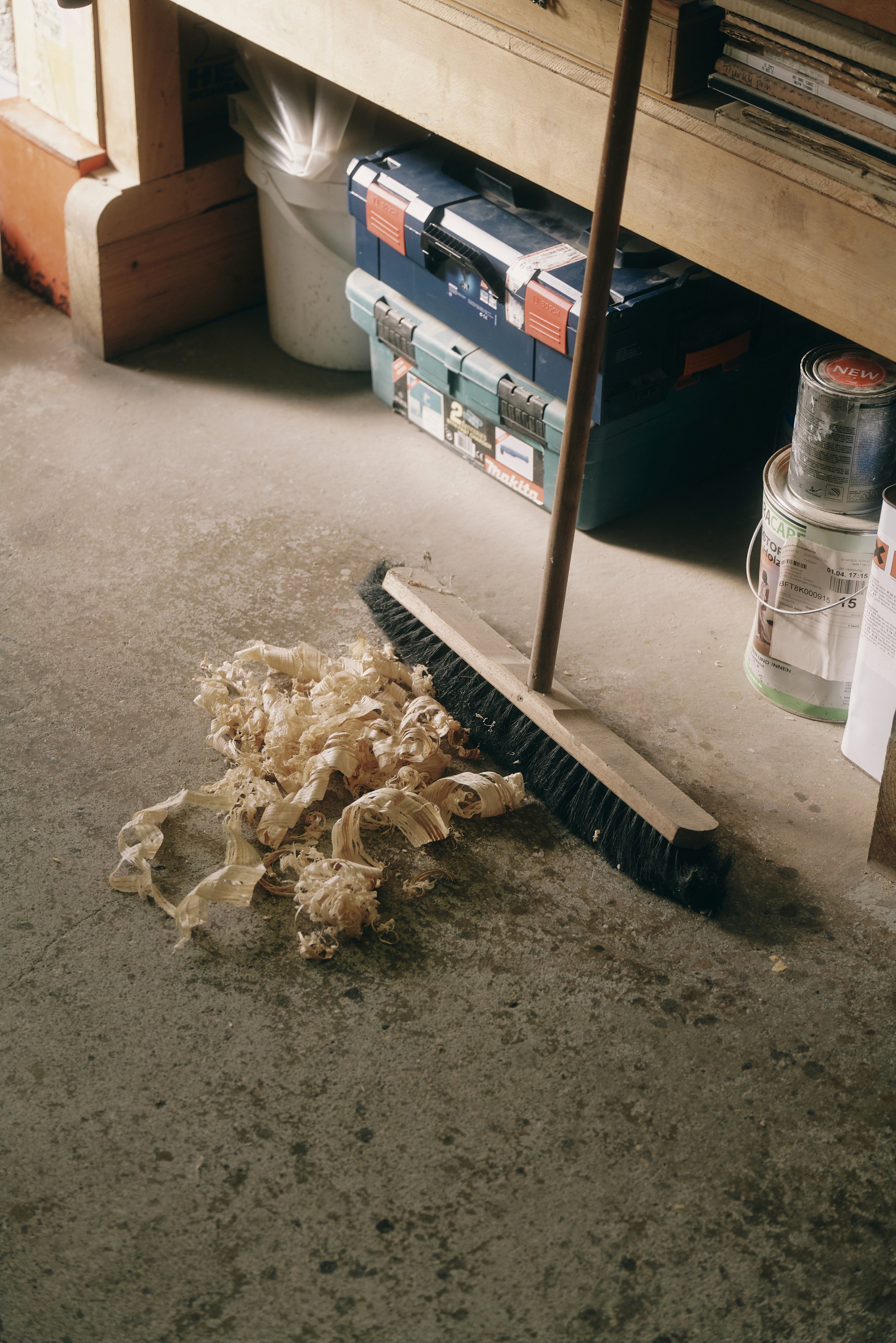 Besenschopf und Sägespäne auf einem Betonboden in einer Werkstatt mit Werkzeugkisten und Farbdosen.