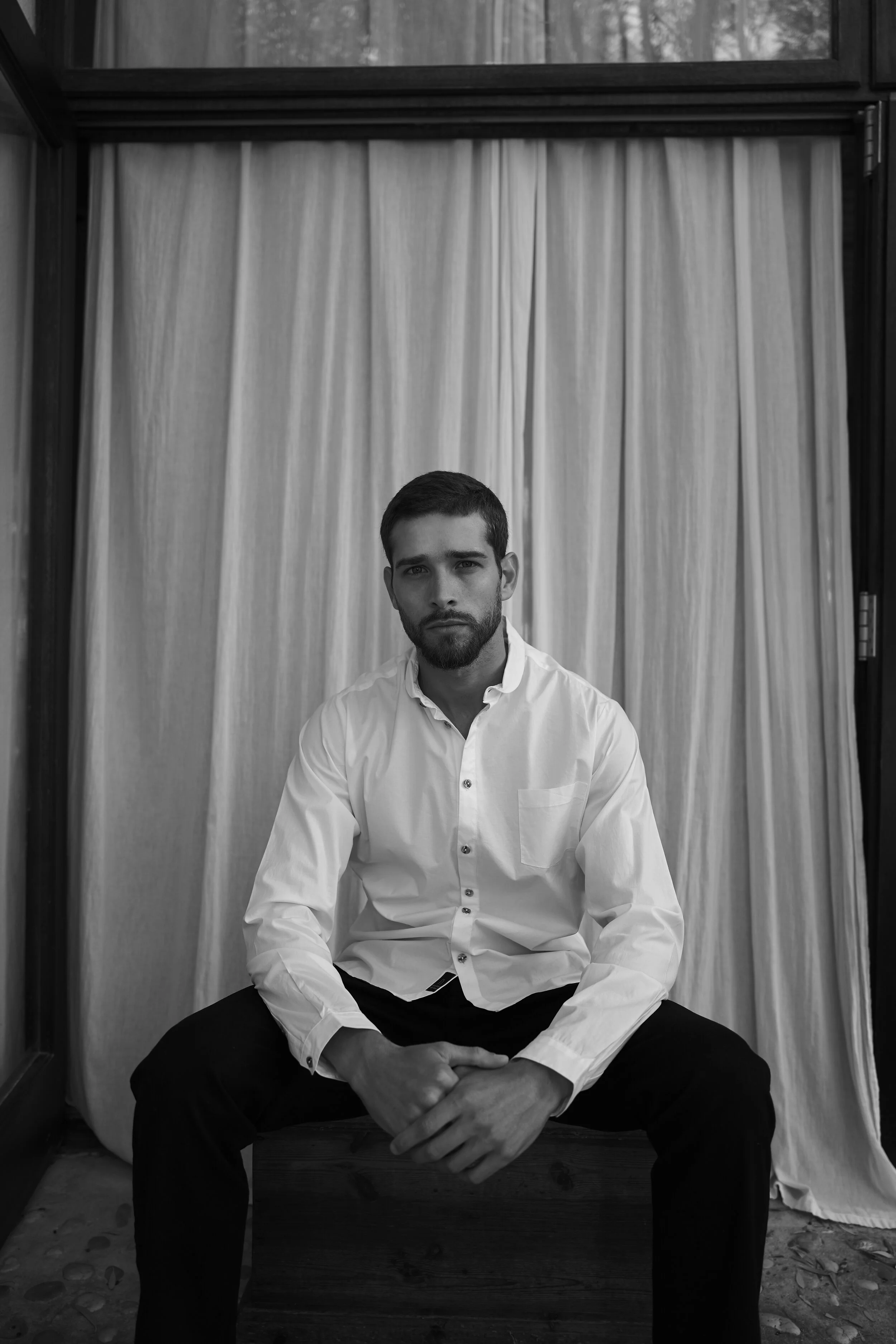 Mann in weißem Hemd sitzt vor Vorhängen.