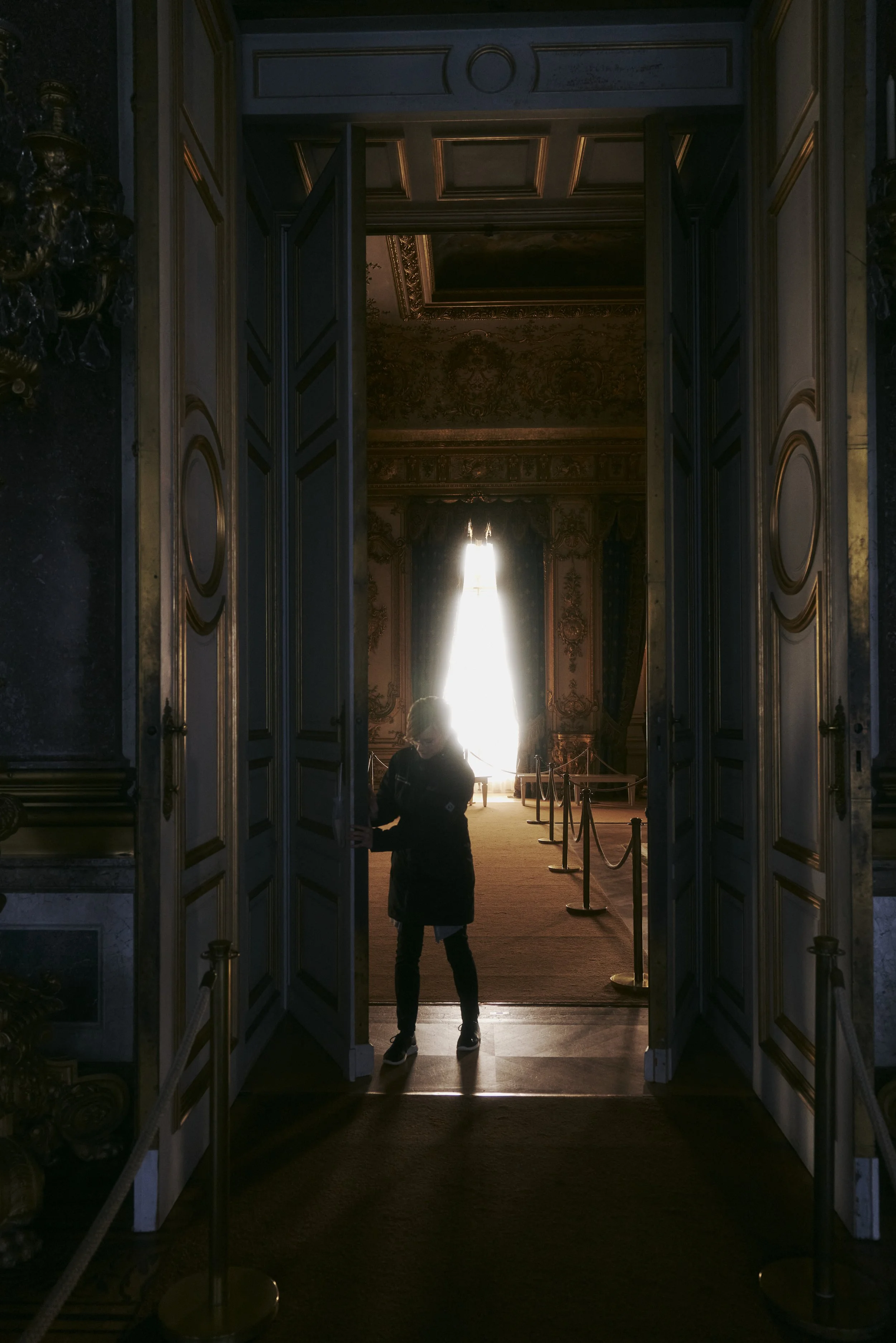 Ein Person öffnet eine große, dekorative Tür zu einem prunkvoll eingerichteten Raum mit glänzendem Tageslicht im Hintergrund.